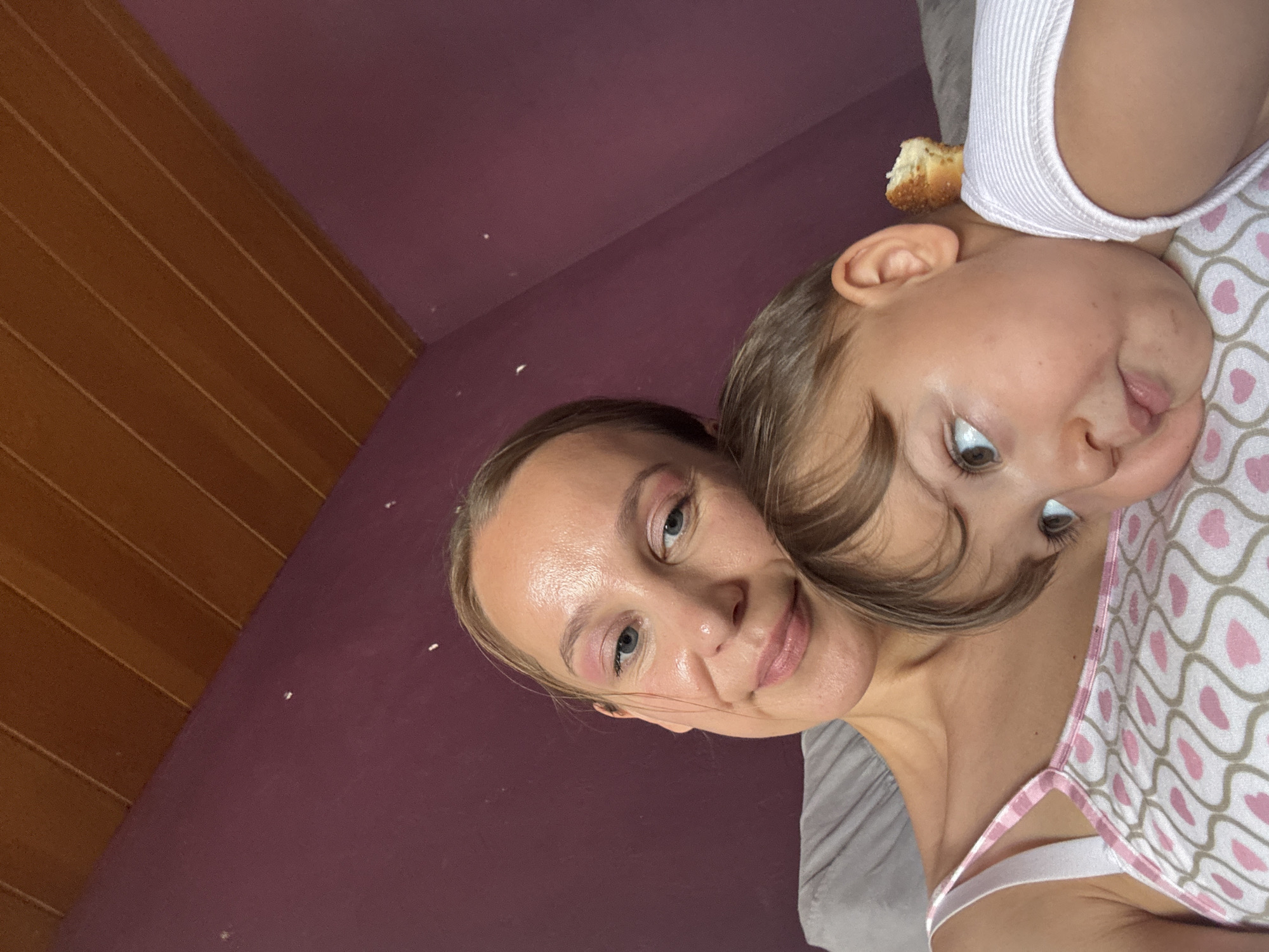 This media file features a close-up, selfie-style portrait of two individuals, an adult and a young child, positioned against a backdrop of a purple wall and wooden paneling. The adult, likely female, is in the foreground, with her head tilted and eyes looking towards the camera. She appears to be wearing light pink eyeshadow and lipstick. The child is positioned behind and slightly above the adult, with their head resting on the adult's shoulder. The child has light brown hair and is looking off to the side, with their mouth slightly open. A small piece of food, possibly bread, is visible on the child's head, near their ear. Both individuals are wearing casual clothing. The lighting suggests it might be daytime, and the indoor setting is indicated by the wall and paneling. The overall mood appears informal and intimate, possibly a moment captured between parent and child. There is no visible text within the frame. The context provided indicates this scene is from Überherrn, Germany.