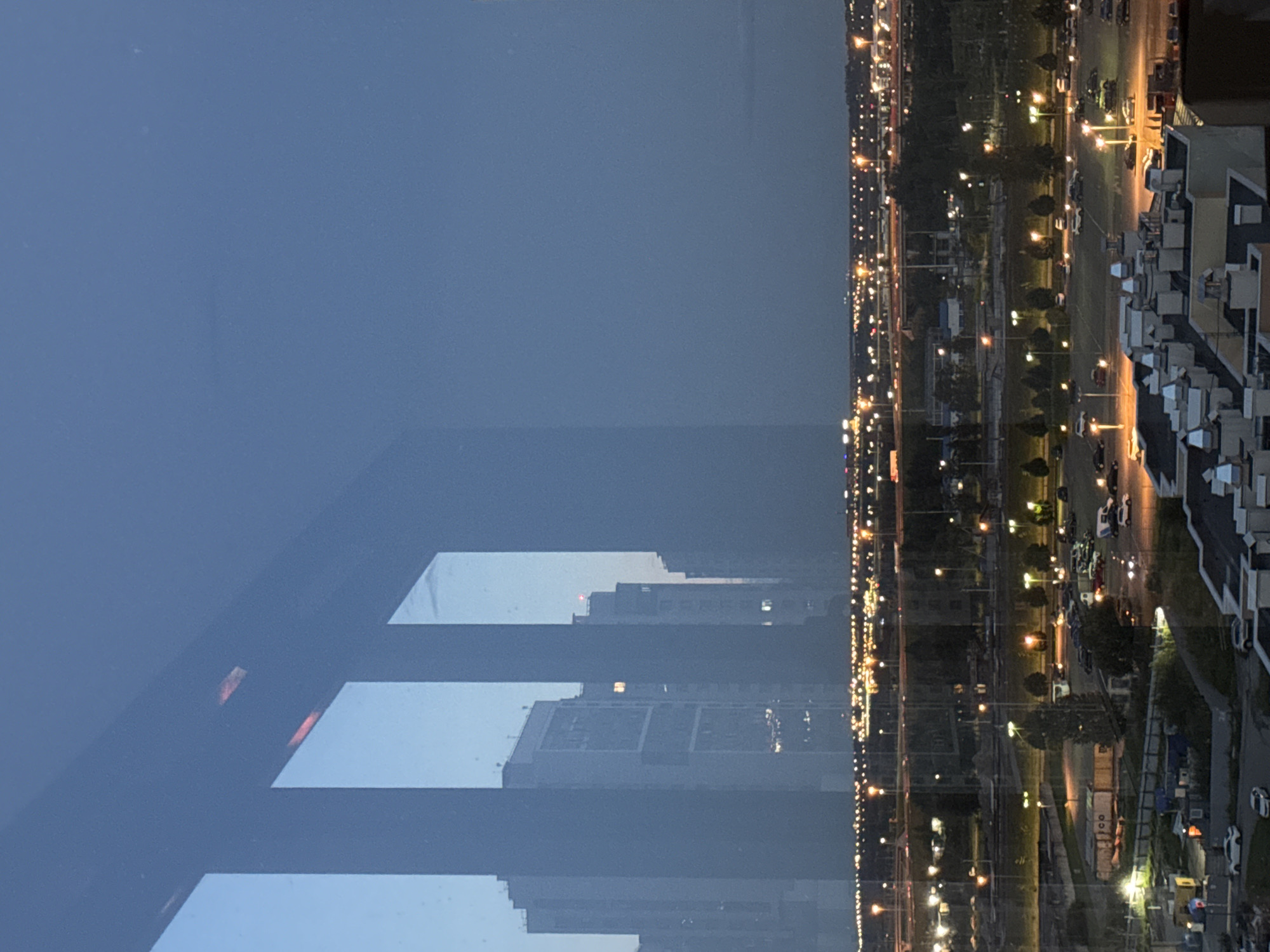 This image depicts a view of a city at dusk, seen through what appears to be a window with reflections. The scene shows a cityscape with several tall buildings, some of which are illuminated. In the foreground, vertical elements of the window frame are visible, casting shadows or creating divisions in the view.
Further out, a wide expanse of lights indicates a developed urban area, possibly a highway or a densely populated district. Numerous streetlights and building lights are visible, creating a shimmering effect across the landscape. Below the distant cityscape, there are darker areas with scattered lights, suggesting a mix of residential and industrial zones, with visible car parks and roads. The sky is a deep blue, characteristic of twilight.
The reflection on the window suggests the presence of an indoor environment, though it is not explicitly shown. No people are clearly discernible in the image. The overall atmosphere is that of a city preparing for the night. Given the context provided, this scene is identified as being in Otradnoye, Russia, a town located within the city of Saint Petersburg.