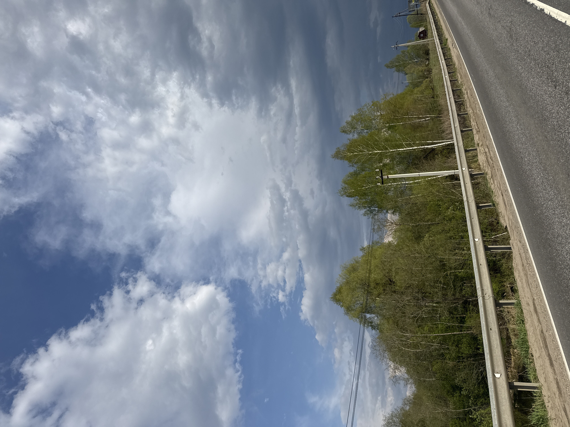 The image shows a view of a road and sky in Razdolye, Russia. The road, visible on the right, has a gray metal guardrail along its edge. Beyond the guardrail is a line of trees with light green leaves, possibly birch trees due to their white trunks. Overhead power lines run alongside the trees. The sky occupies a large portion of the image and exhibits a mix of dark gray storm clouds and patches of bright blue sky with white, puffy clouds. The weather appears to be transitioning, with elements of both clear and overcast conditions.