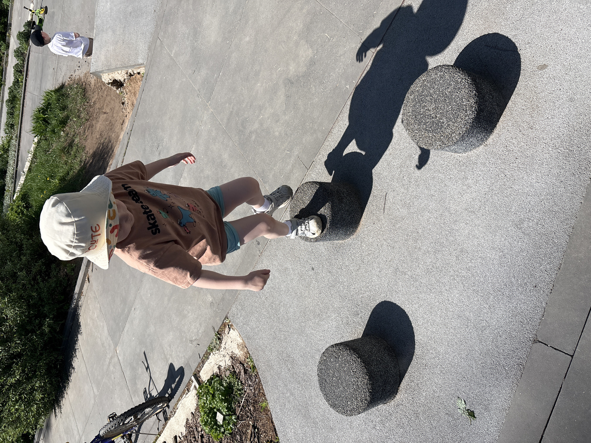 A child wearing a tan t-shirt and a white bucket hat with the word "cute" printed on it is stepping on a dark gray, cylindrical, textured object. Two other similar objects are nearby. The child is casting a shadow. A person wearing a white shirt is visible in the background near a landscaped area. The surface is concrete, divided into sections. A bicycle lies on the ground near a plant bed. The photograph was taken outdoors in daylight. I am unable to establish the location as Izhevsk, Russia.