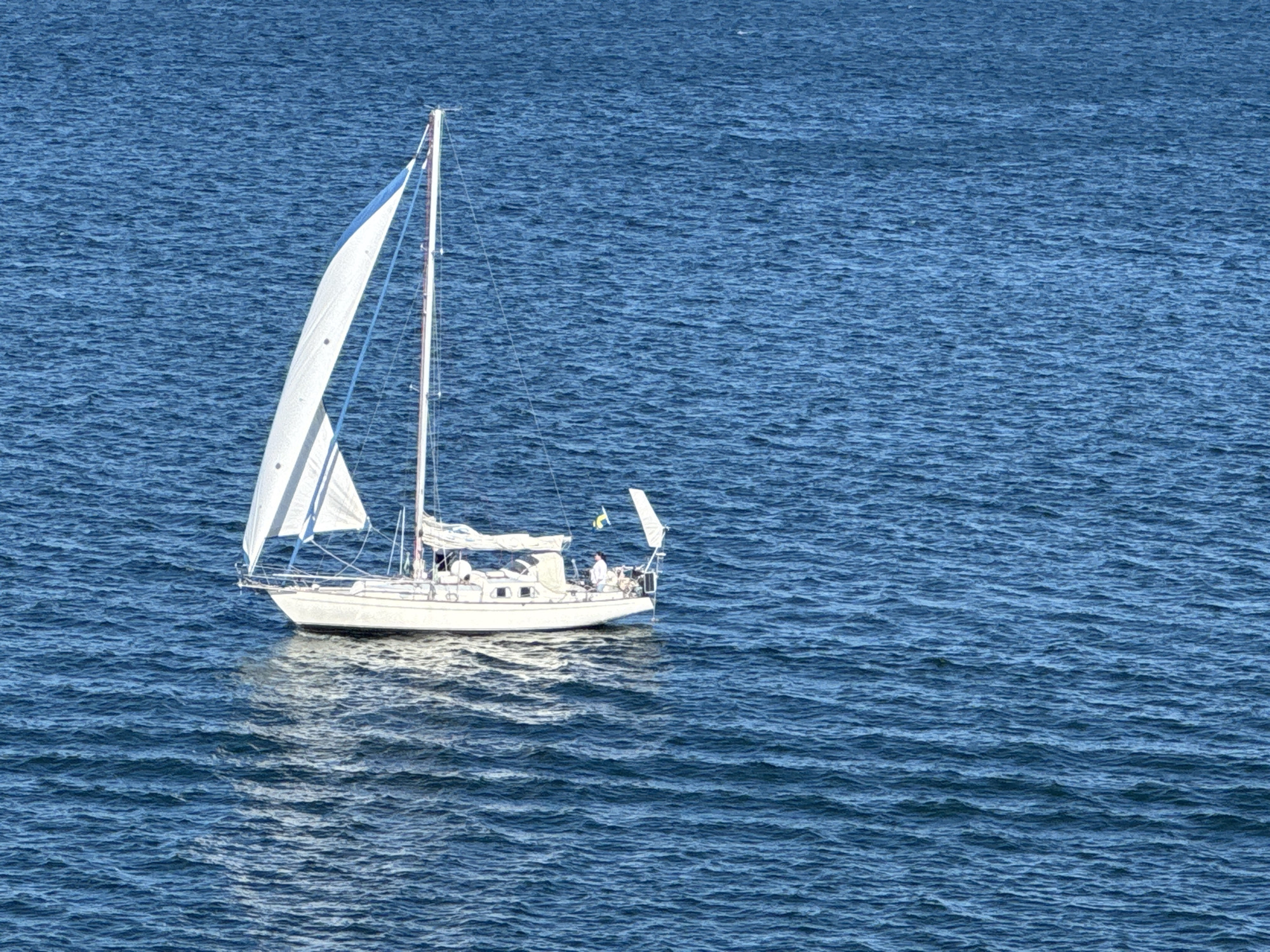A white sailboat with a blue stripe on its sail is navigating through choppy blue water. The boat has a white hull and a mast with rigging. Two people are visible on the deck of the sailboat. The person at the helm appears to be wearing a white shirt, and another individual is standing near the stern. A small yellow and blue flag is attached to the back of the boat. The water exhibits ripples and reflects the sunlight, creating a textured surface. This scene is likely taking place in the waters surrounding Karlskrona, Sweden, a city known for its naval heritage and archipelago.