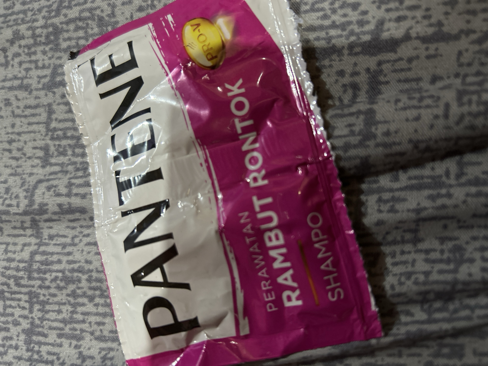 A Pantene shampoo sachet is photographed against a grey textured fabric. The sachet has a white top portion with the brand name "PANTENE" printed in large black letters. Below the brand name, a pink section contains text that reads "PERAWATAN RAMBUT RONTOK" and "SHAMPO". A yellow oval with "PRO-V" is visible on the right side of the pink section. The sachet is lying flat on the fabric, which appears to be a bedspread or similar textile with a repeating pattern. The lighting suggests an indoor environment. There is no information or visual evidence in the image to indicate the location of Depok, Indonesia.