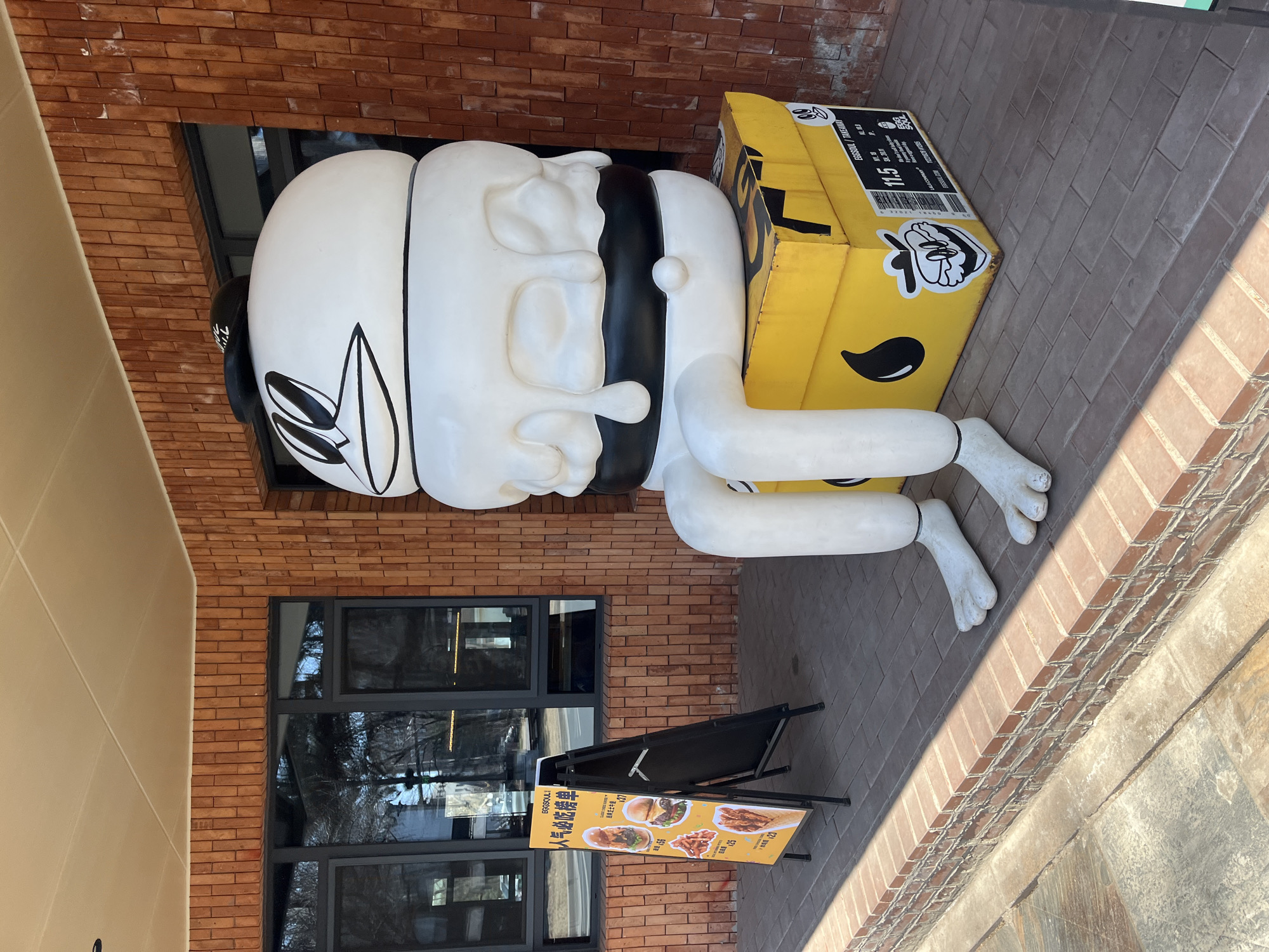 A full, medium-shot photograph shows a large sculpture of a man carrying a yellow box on his back in Changchun City, China. The sculpture is located outside of a building with red brick and a window.
The sculpture is white with black accents. The man has a large, round head, bulbous nose and closed eyes, and a black neckerchief. The man's legs and feet are positioned on the ground. He is carrying a yellow box on his back that is labelled with food information.
A black, triangular sign with a yellow background and text and photos is on the ground in front of the sculpture. The background includes the building, pavement, and sunlight.