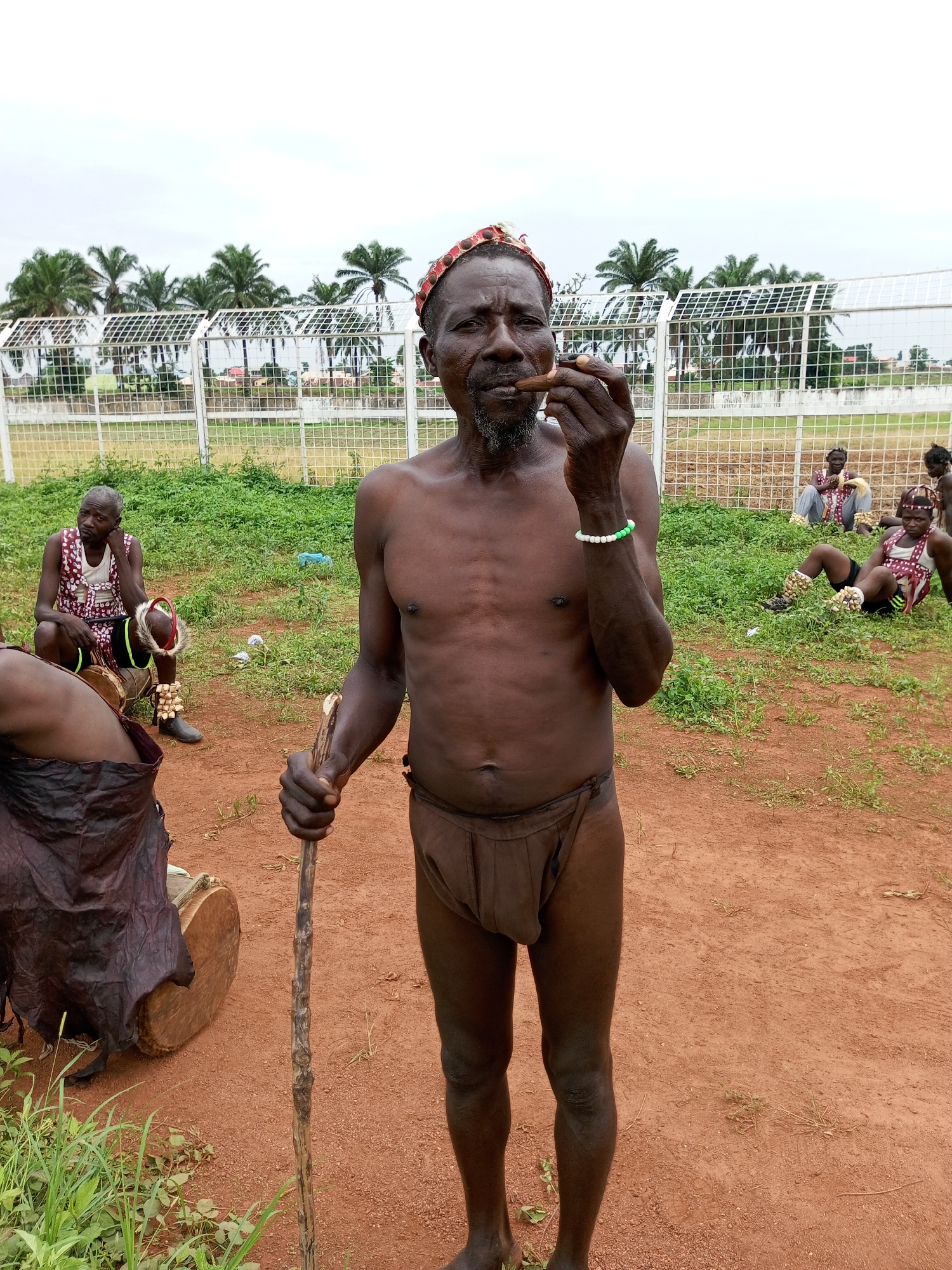 The image captures a scene in Takau, Nigeria. A shirtless, elderly man wearing a loincloth and a patterned head covering stands in the foreground. He holds a wooden staff and smokes a cigar. The ground is a mix of red dirt and sparse green vegetation. Behind him, a group of people are either sitting or lying on the grass. One man is seated on a drum, wearing a patterned vest and leg ornaments. In the background, a white fence and palm trees are visible against a cloudy sky.