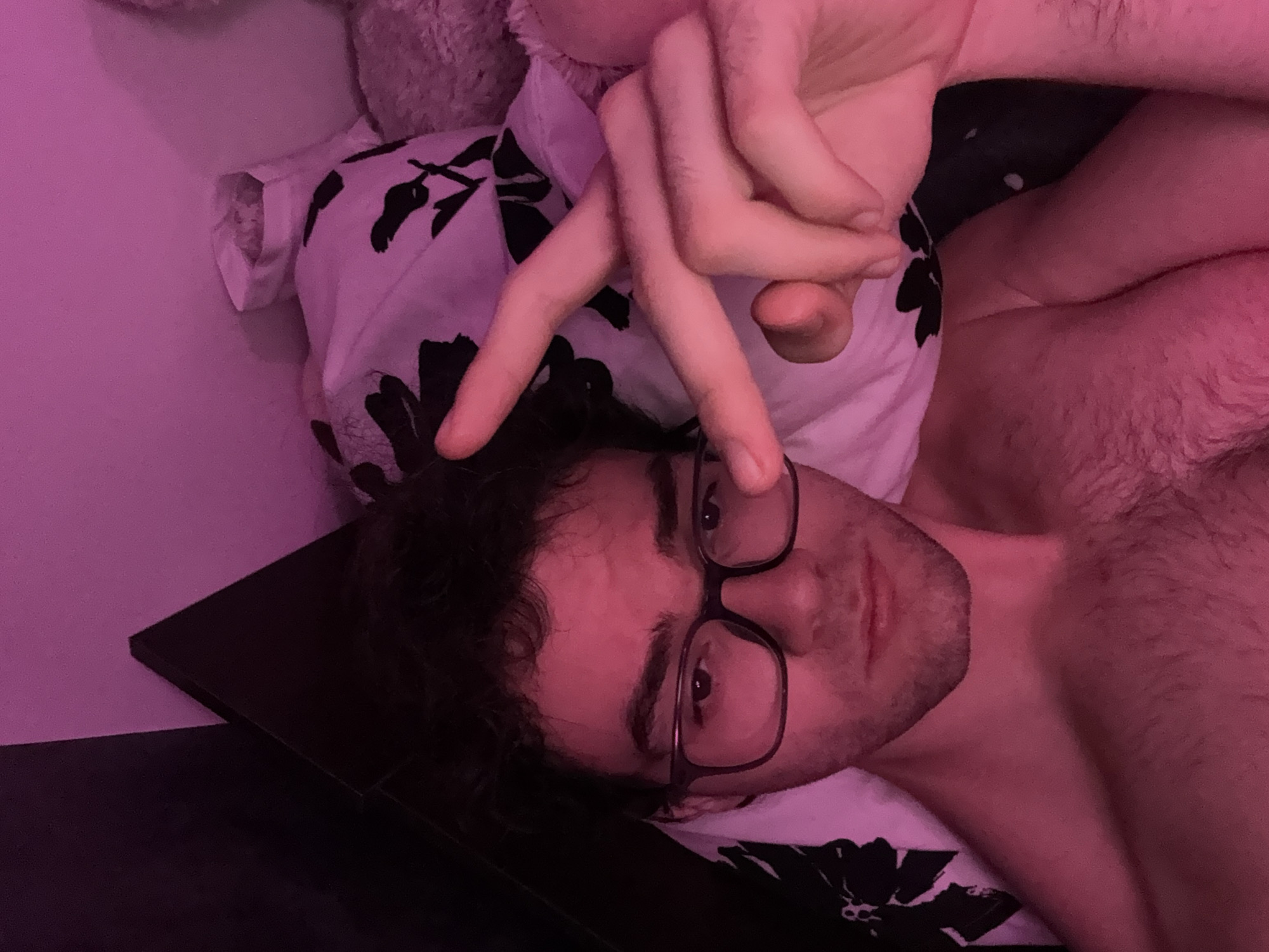 The image is a selfie of a person lying in bed. The person is wearing glasses and has curly, dark hair. Their right hand is raised, with two fingers extended in a peace sign. The background shows a pillow with a black and white floral pattern and a textured surface that appears to be a blanket or stuffed animal. The scene is illuminated by pink lighting, possibly from a light bulb or ambient light. There is no discernible information about the location within the city of Blagoevgrad, Bulgaria, as the image is confined to the personal space of the individual.