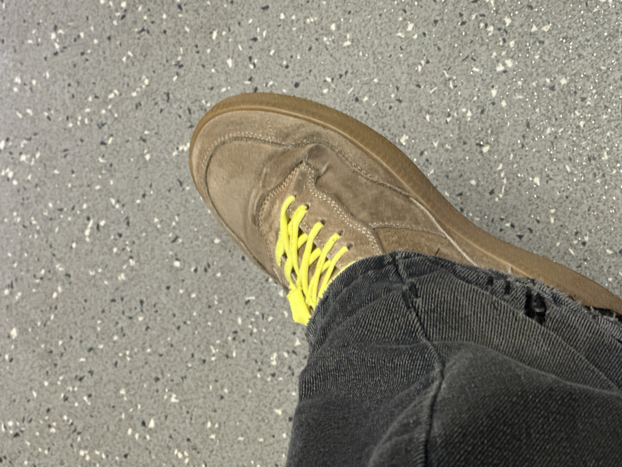 The image shows a person's foot wearing a tan suede sneaker with bright yellow laces. The person is also wearing dark grey denim jeans. The background is a speckled grey floor, likely a public space. It is impossible to determine the exact location as Moscow, Russia based on the visible details alone.