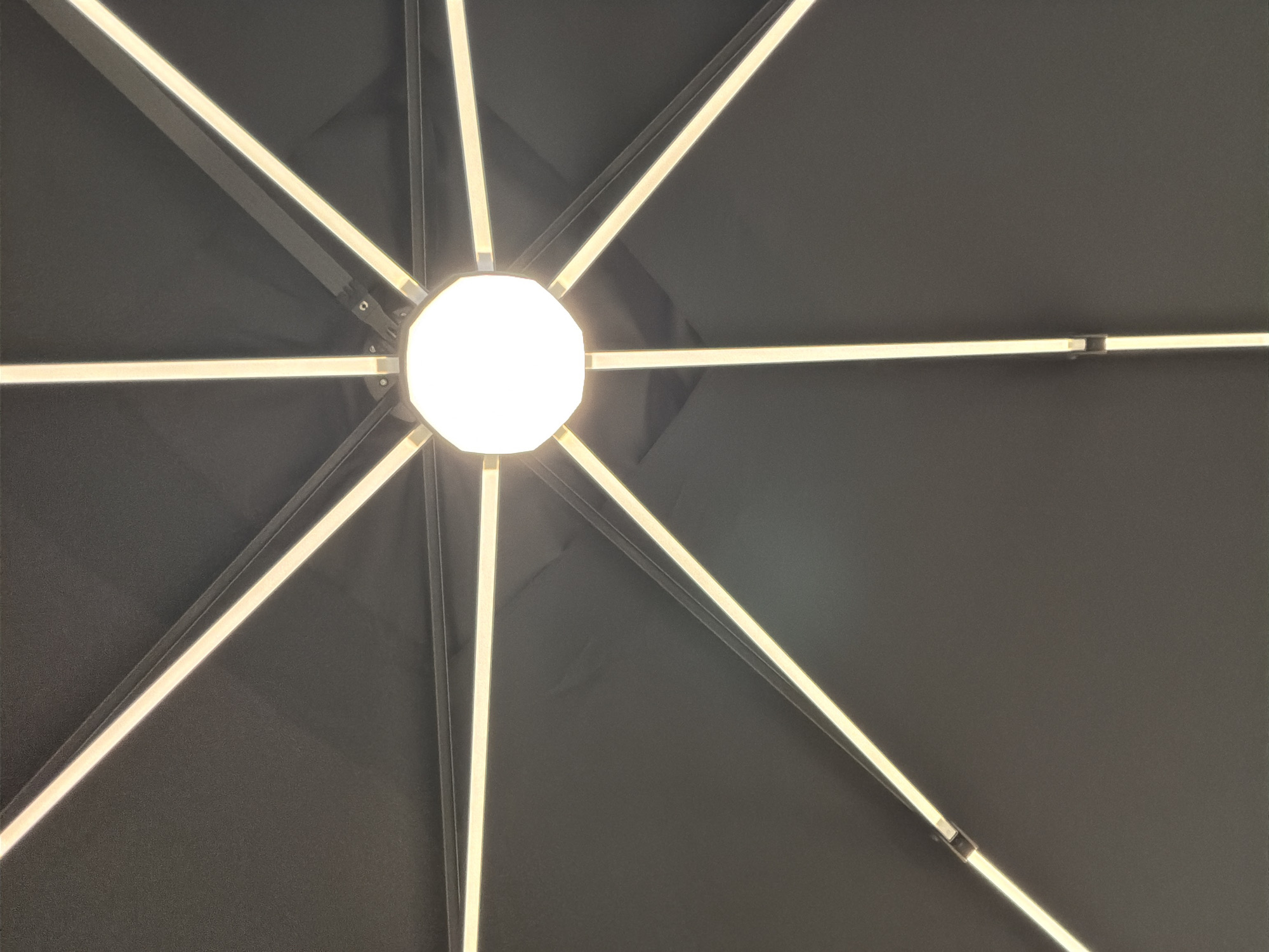 The image is a close-up, upward shot of the underside of an umbrella, likely in a city such as Ipswich, United Kingdom. The umbrella's canopy is a dark gray or black fabric stretched taut over a frame of eight radiating metal supports. Integrated into each support is a strip of white LED lighting, illuminating the dark canopy with a bright, linear glow. At the center of the umbrella, where the supports converge, is a large, octagonal light fixture, also emitting a bright, white light, creating a starburst pattern of illumination against the dark fabric. The overall effect is a modern and functional design for providing shade and light, possibly in an outdoor dining or seating area.