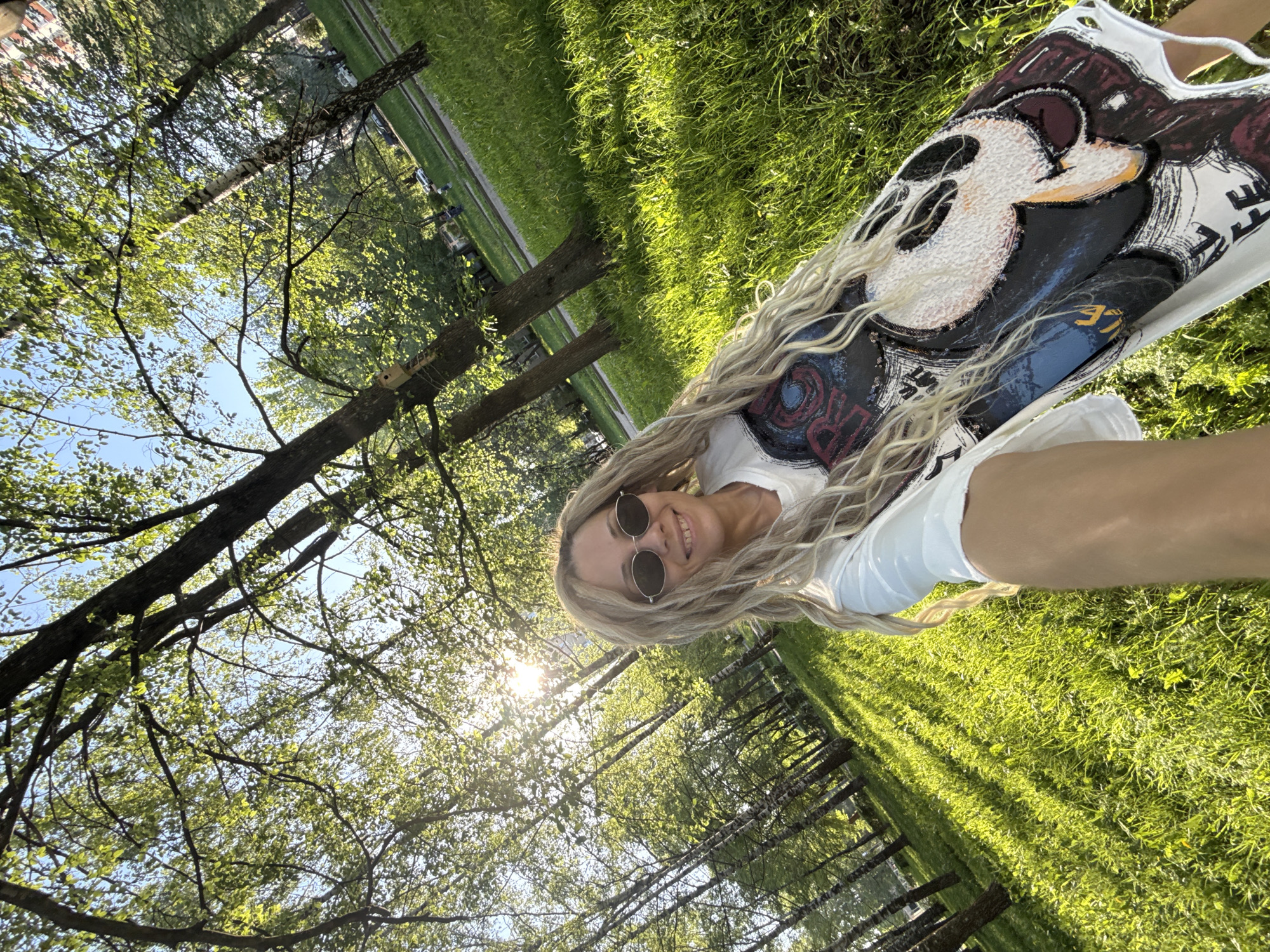 The image shows a woman with blonde hair wearing sunglasses and a white T-shirt with a Mickey Mouse design. She is smiling and appears to be taking a selfie. The background consists of trees and green grass, suggesting a park setting. The sun is shining through the trees, creating a bright, dappled light effect. The location is Rusanovo, Russia.