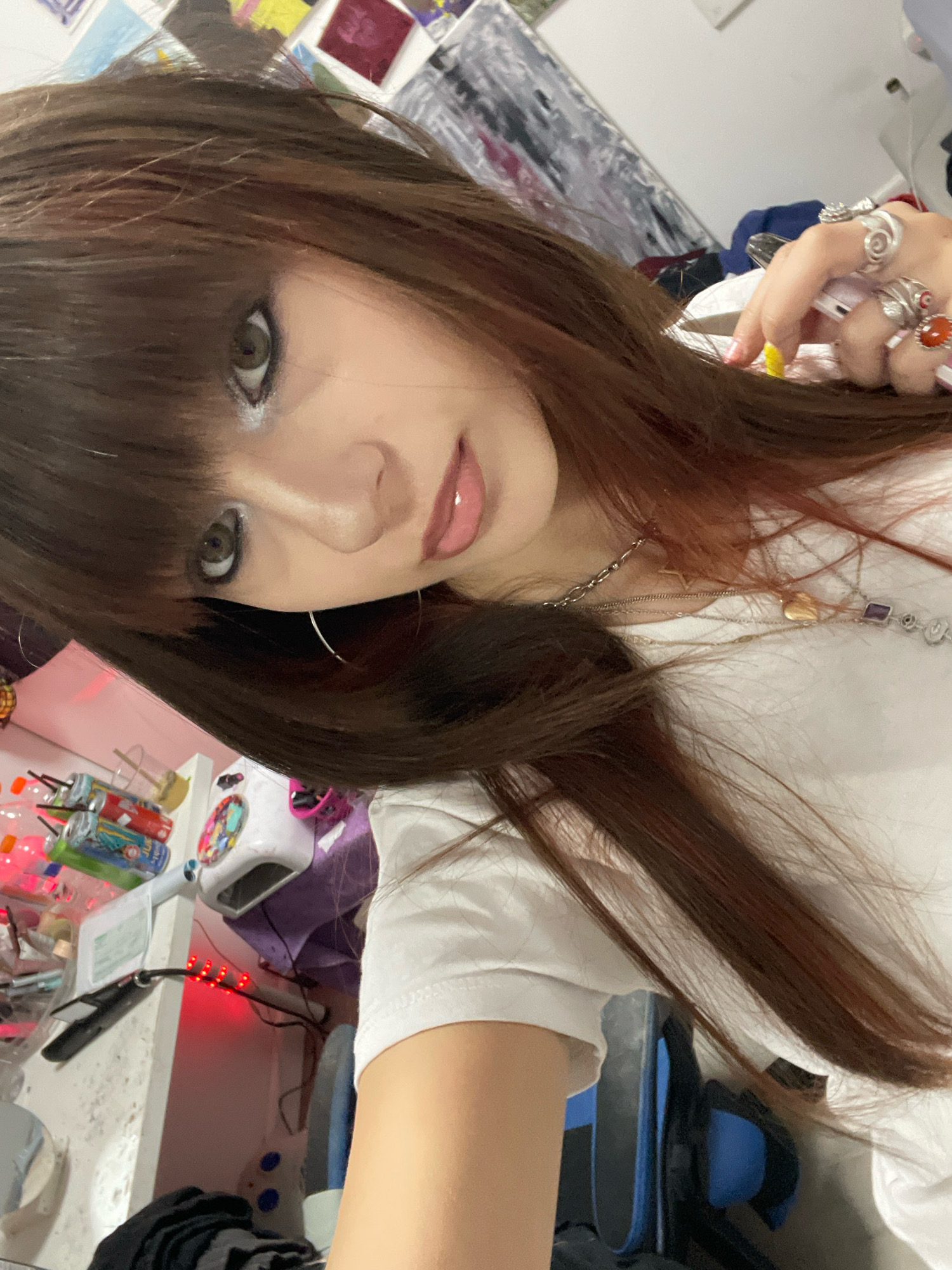 The image, taken in what appears to be a room in Raanana, Israel, is a close-up, tilted selfie of a young person with long, dark brown hair. The subject is wearing a white t-shirt and has visible makeup, including eyeshadow and lip gloss. Several rings adorn their right hand, including one with a prominent orange stone and another with an evil eye design. A silver necklace with a heart pendant and a purple gemstone is also visible. In the background, various objects are scattered, suggesting a cluttered desk or workspace. These include what appear to be small bottles or containers, a white device with red LED lights, and what might be art supplies or personal items. The overall impression is that of a casual, personal moment captured in a domestic setting.