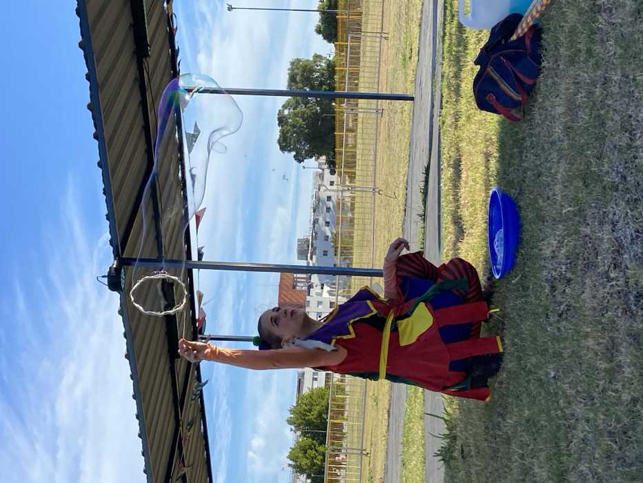 A performer in colorful garb stands outdoors, making a giant bubble. She holds a looped wand, drawing a shimmering, elongated bubble from it that drifts upward. She wears a red, purple, and yellow patchwork dress, and orange arm coverings. A blue plastic bowl and a striped bag sit on the grass nearby. The scene is partially shaded by a structure overhead, with a clear sky visible beyond. Fencing and buildings form a distant backdrop.
