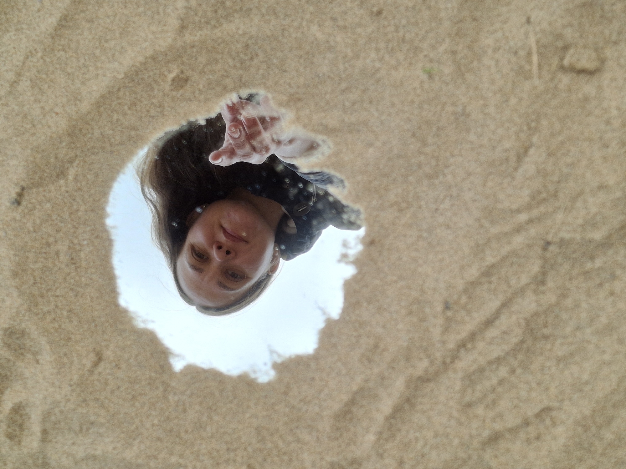 The image depicts a woman's face and hand as seen through a hole in the sand. Her hand extends towards the viewer. The background visible through the hole is a cloudy sky. The scene appears to be on a beach or sandy area. No further information is available about the location in the city of Chaikovsky, Russia, or the actions or interactions of the person or objects.