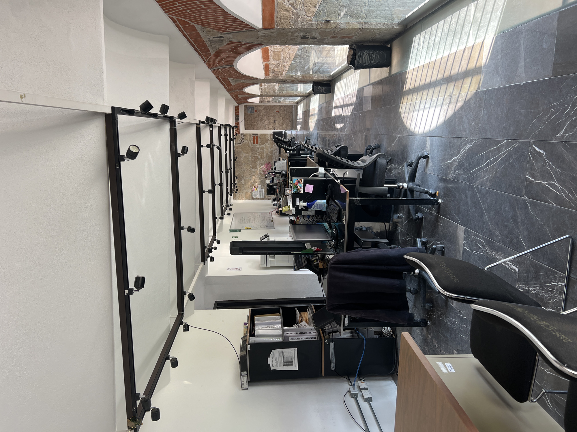 The image depicts a long, narrow office space in Puebla, Mexico, characterized by a blend of modern and traditional architectural elements. The office features a row of workstations along one side, each equipped with a computer, monitor, and office chair. The opposing wall consists of white panels, possibly display boards, illuminated by numerous small spotlights mounted on dark frames. The workstations are cluttered with office supplies and equipment. Overhead, the ceiling transitions from a modern white section to a series of arched brick structures, evoking a historic feel. Natural light streams in from a window along the workstation side, casting shadows on the dark gray, marble-like tiled floor. The scene appears unoccupied.