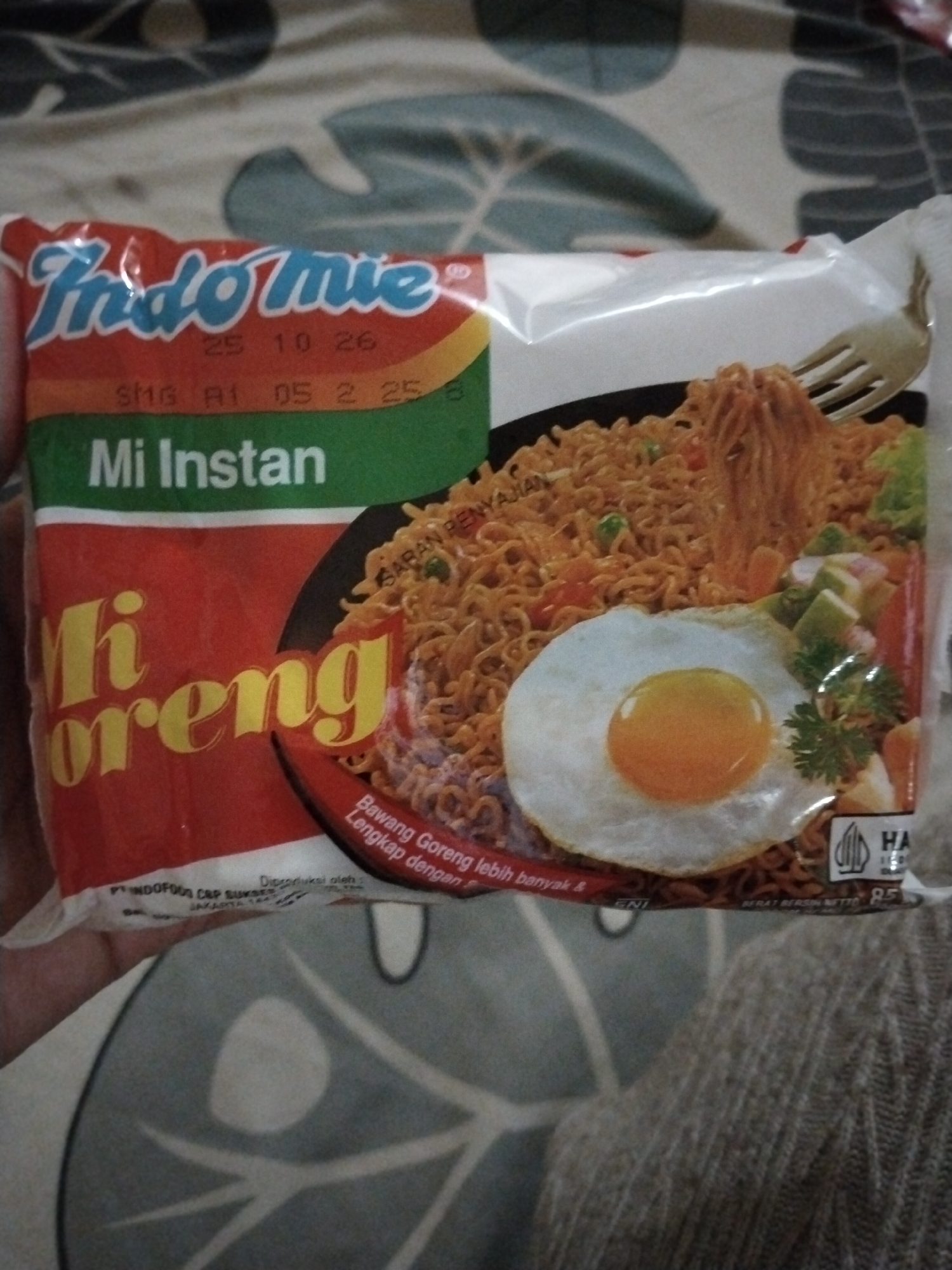 The image shows a package of Indomie Mi Goreng instant noodles, a popular food product from Indonesia. The packaging is predominantly red with the brand name "Indomie" in blue lettering at the top. The product name "Mi Instan" (instant noodles) is in white text on a green background, and "Mi Goreng" (fried noodles) is in large yellow lettering. A picture of the prepared dish, featuring noodles, an egg, and vegetables, is visible on the right side of the packaging. The package is placed on a textured fabric surface, possibly a bedsheet or blanket, with a green and white patterned design visible in the background. The image is taken from a slightly angled, close-up perspective. There is no indication of any specific location within Kecamatan Pati, Indonesia, or any people in the image.