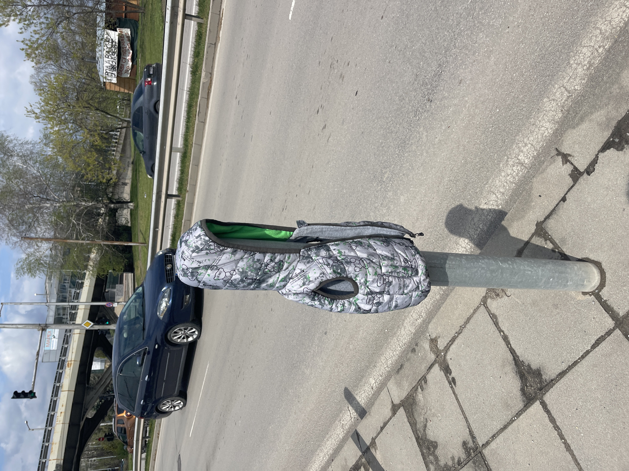 A gray and white patterned dog coat is hanging over a metal pole. The coat has green lining and a gray zipper. The pole is located on a sidewalk made of cracked, square, concrete tiles. A street runs alongside the sidewalk. Cars are parked and driving on the street. Buildings and trees are visible in the background, as well as an elevated bridge. The image was taken during the day in Sofia, Bulgaria. A "Mapet" sign is visible on the elevated bridge.