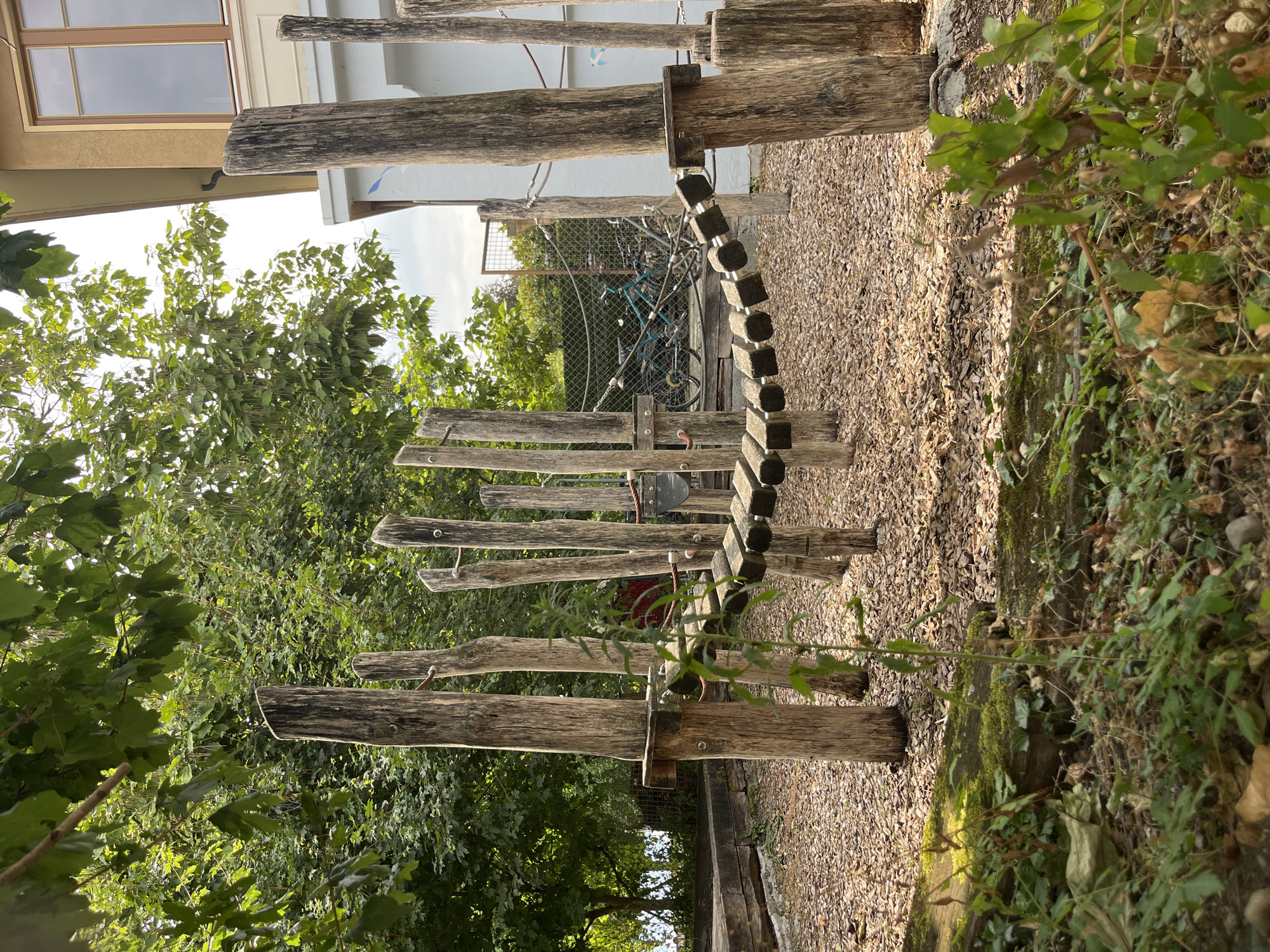 The image depicts a natural playground constructed with wooden logs and ropes, set against a backdrop of lush green trees and a building. The playground features a suspended bridge made of wooden planks and rope, supported by vertical wooden posts. The ground is covered with wood chips. In the background, several bicycles are visible behind a fence. The scene suggests an outdoor recreational area, possibly a park or a schoolyard, located in Vevey, Switzerland. The lighting indicates it is daytime, with natural sunlight illuminating the scene. There are no people visible in the image.
