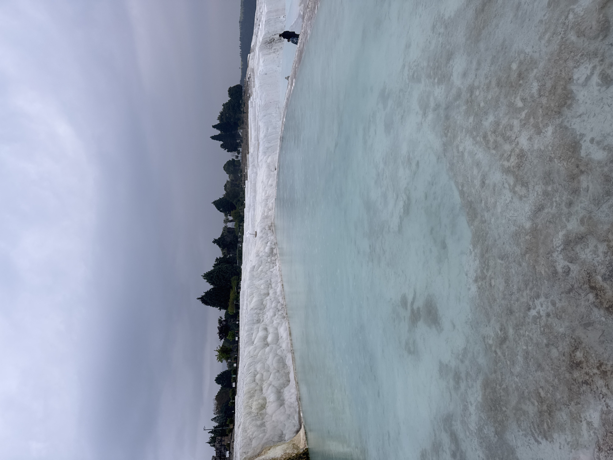 This image shows a view of the travertine terraces of Pamukkale, Turkey. The foreground features a large pool of light blue water with grey textured sediment on its edges. The white, bubble-like travertine formations border the pool. A line of trees is visible behind the formations, leading up to a cloudy, overcast sky. A single figure can be seen standing near the upper edge of the travertine. The image is angled, emphasizing the slope of the terraces.