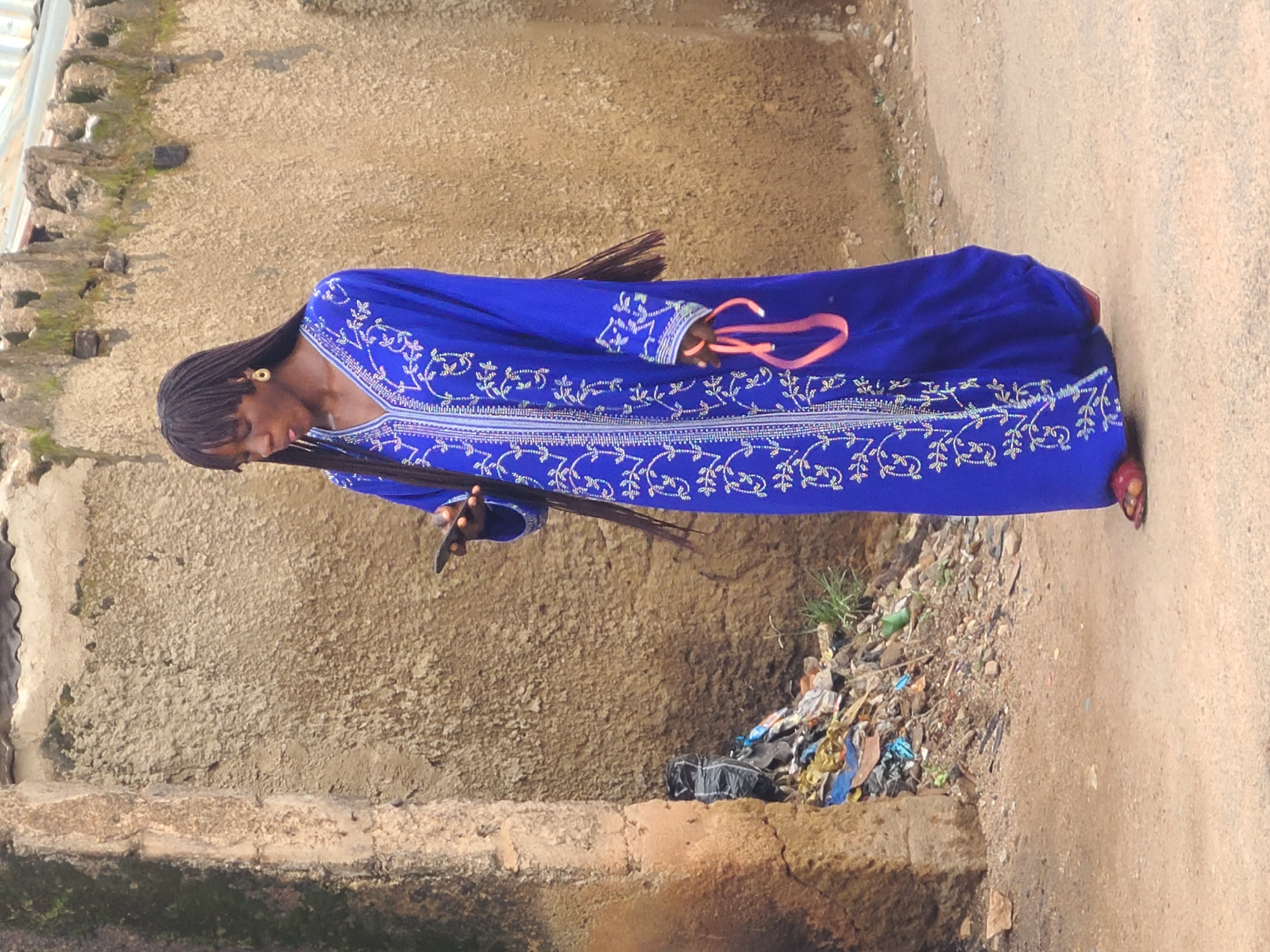 A young Black woman, with long dark braided hair reaching past her shoulders, is standing in what appears to be a dusty, undeveloped area. She is wearing a long, royal blue, floor-length dress adorned with intricate white embroidery and silver beadwork. The dress has a V-neckline and long sleeves. In her right hand, she holds a pink, ribbon-like object, possibly a hair tie or decorative sash. Her left hand is holding a black smartphone up to her face, as if she is looking at it or using it. She is wearing red slip-on sandals.
Behind her is a rough, textured, beige-colored wall, likely made of mud or concrete, with patches of green moss growing on it. To her left, at the base of a rocky outcrop, is a pile of assorted trash, including plastic bags, bottles, and other debris, indicating a less-than-sanitary environment. The ground is a mixture of dirt and gravel. The overall scene suggests an outdoor setting in a place with limited infrastructure. This environment is consistent with areas found in Jos, Nigeria.
