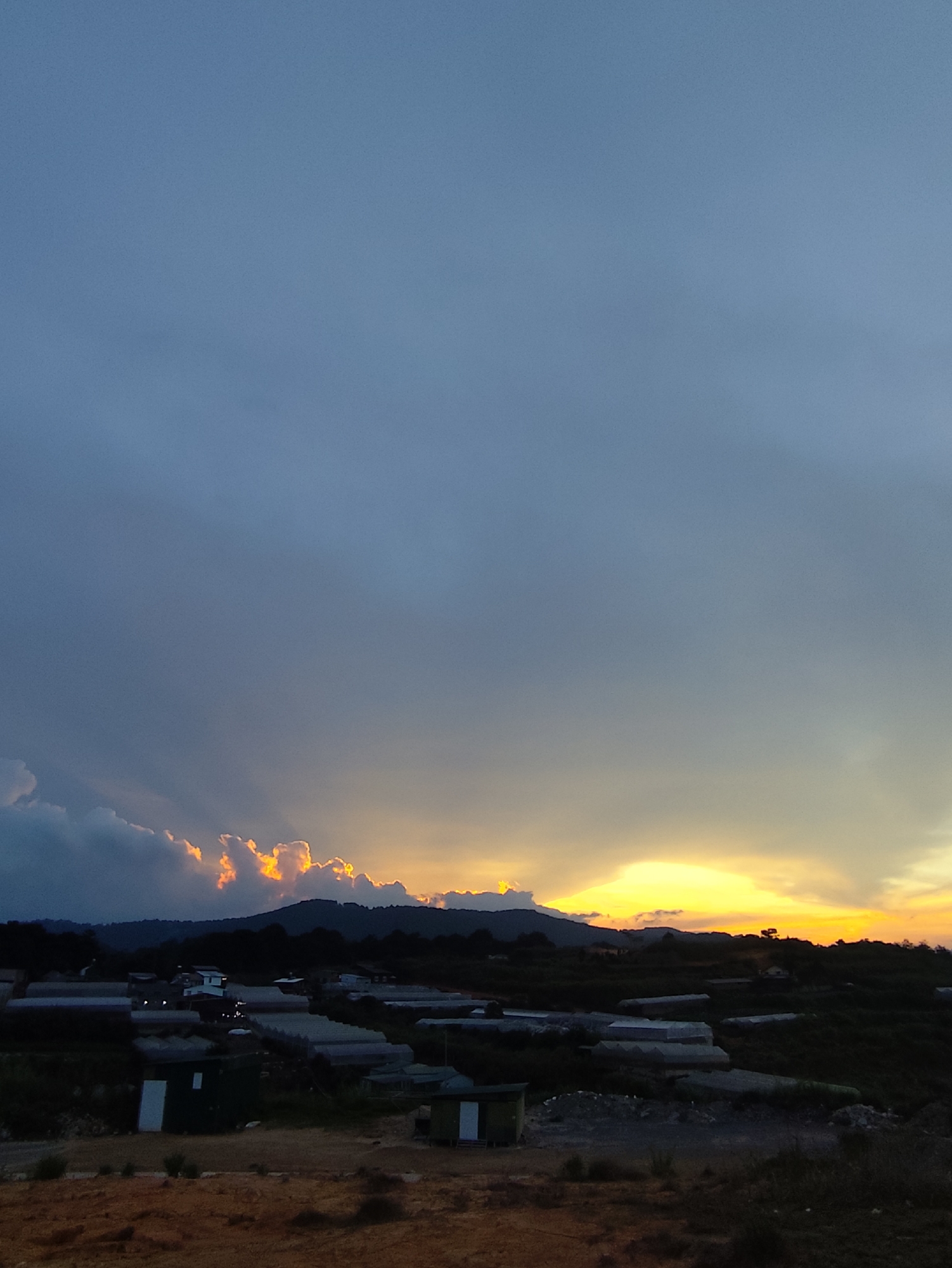 The image captures a sunset over what appears to be an agricultural area, possibly in the vicinity of Da Lat, Vietnam. In the foreground, there is a dirt ground area, with a scattering of small buildings, perhaps farm structures, and a significant presence of what seem to be greenhouse structures. These structures are rectangular, low to the ground, and covered in a semi-transparent material, possibly plastic or glass.
In the middle ground, the terrain rises towards a series of hills, silhouetted against the colorful sky. The sky itself is a mix of shades, with a gradient from a dark blue above to a bright orange and yellow near the horizon, indicating a sunset. The sunlight is filtering through some clouds, creating a display of light rays. There are no people visible in the image. The overall environment is rural or semi-rural, with an emphasis on agricultural activities.
