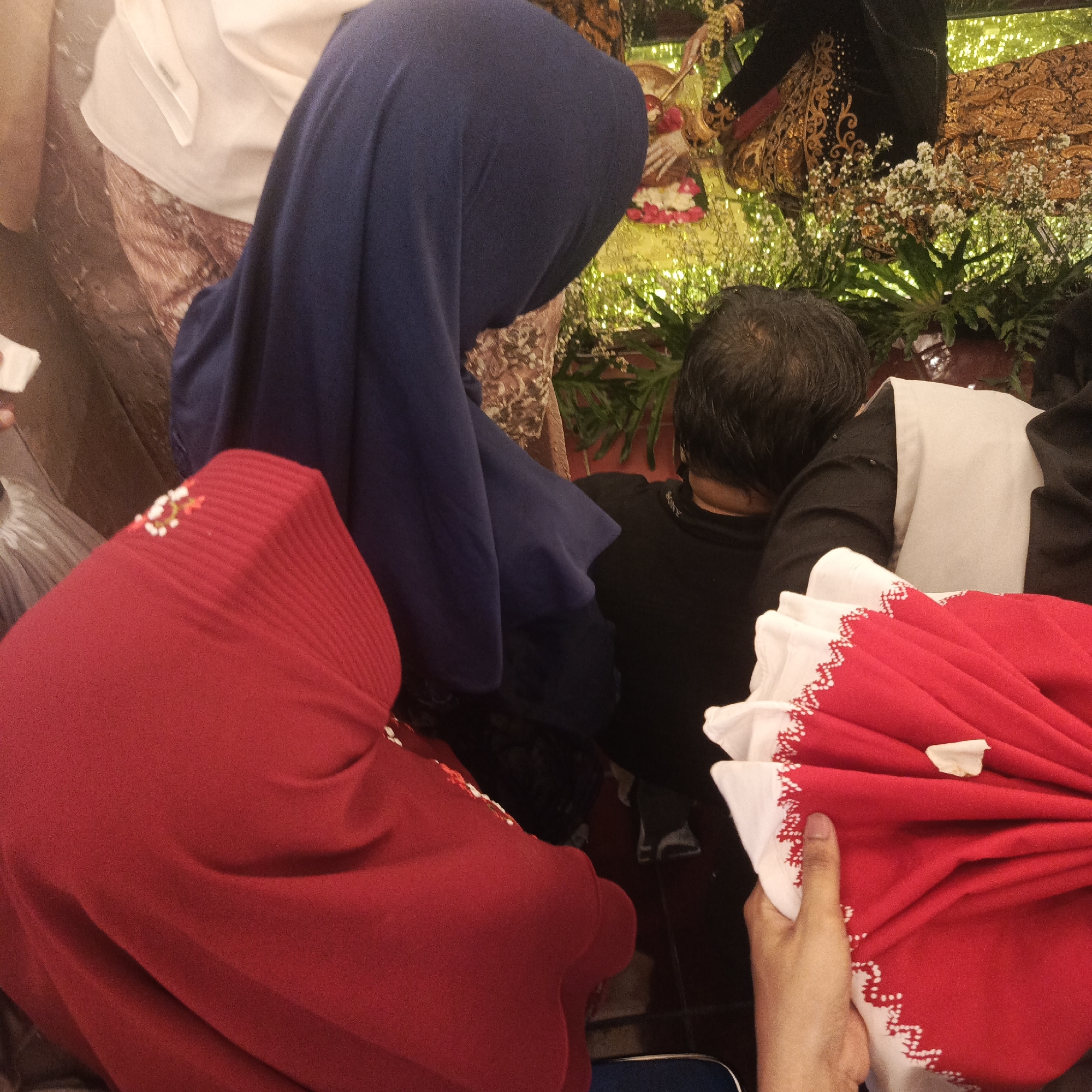 The image, taken indoors, depicts a gathering of people, likely at an event in South Jakarta, Indonesia. In the foreground, two individuals are prominently visible. One person is wearing a bright red headscarf with a beaded embellishment. Another person is wearing a dark blue headscarf. Their bodies are angled towards the center of the frame, suggesting they are interacting with something or someone just out of view. In the middle ground, a person with dark hair is facing away from the camera, wearing a black shirt and a light-colored vest. To the right, a person in a black shirt with ornate gold and black fabric is visible, suggesting traditional attire. A hand is seen holding a folded red fabric with white decorative trim. The background is blurred but suggests a decorated space with foliage and white floral arrangements, possibly indicative of a festive occasion such as a wedding or celebration. The lighting appears to be artificial and warm.