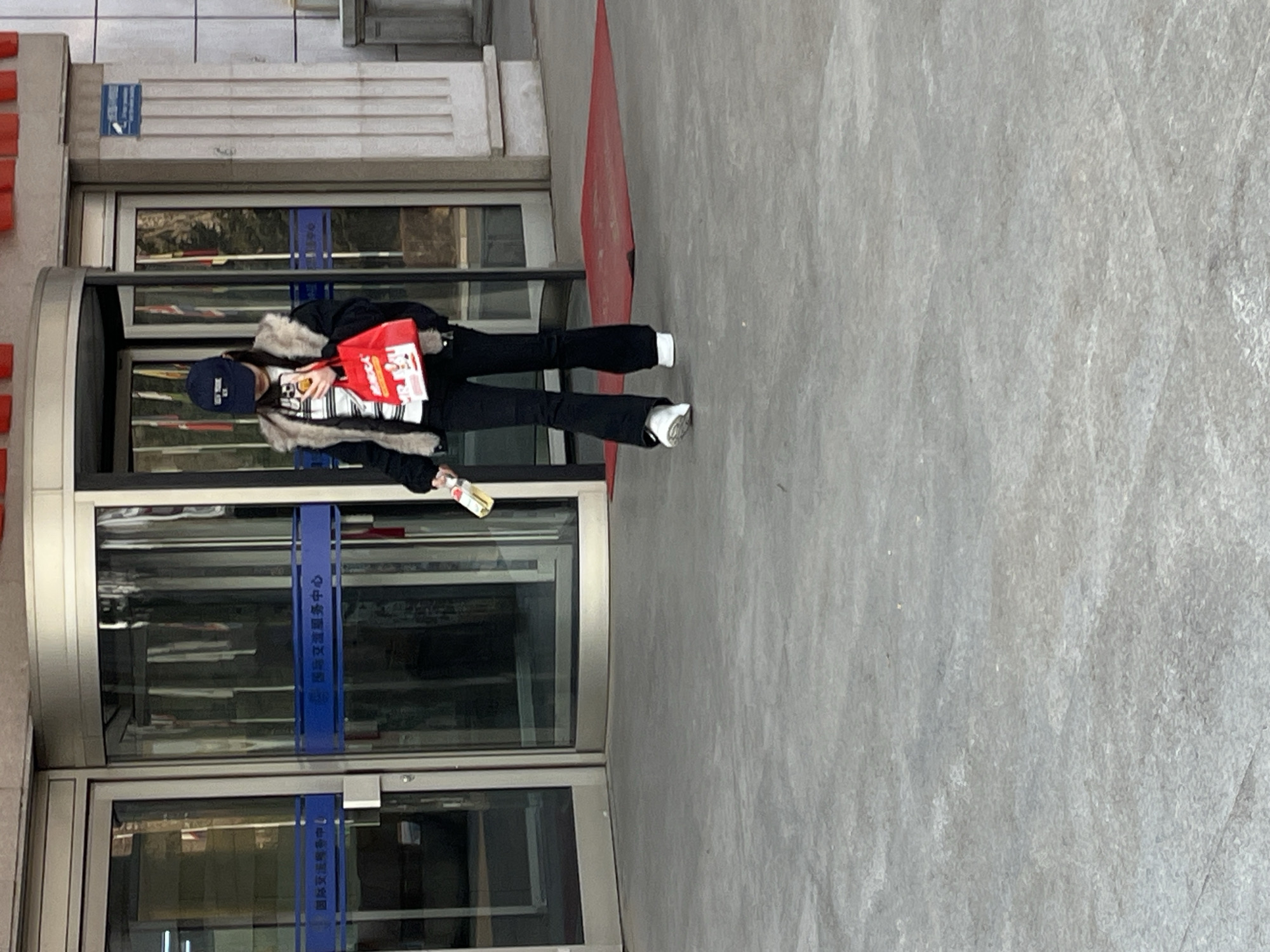 The image shows an individual exiting the Changchun International Exchange Service Center, located at No. 5626 Renmin Street, Chaoyang District, Changchun City, Jilin Province, China. The person, appearing female, is positioned in the foreground, walking away from the building's main entrance. She is wearing a navy blue baseball cap, a dark jacket with a light-colored, possibly fur, collar, a white and black striped long-sleeve shirt, black flared trousers, and white athletic shoes. In her left hand, she carries a red shopping bag with white text and a graphic. In her right hand, she holds a smartphone with a yellow case and a clear plastic bottle containing a light yellow liquid.
The building entrance features a large glass revolving door centrally located, flanked by two standard glass doors. Blue horizontal bands across the glass panels display the Chinese characters "国际交流服务中心" (International Exchange Service Center). The building's facade is light-colored, exhibiting vertical fluting or pillar-like structures to the right of the entrance and red architectural elements above the doors. A small blue sign is affixed to the facade to the right. A red floor mat is visible on the gray concrete ground directly outside the revolving door. Reflections of flags and interior elements are present in the glass doors.