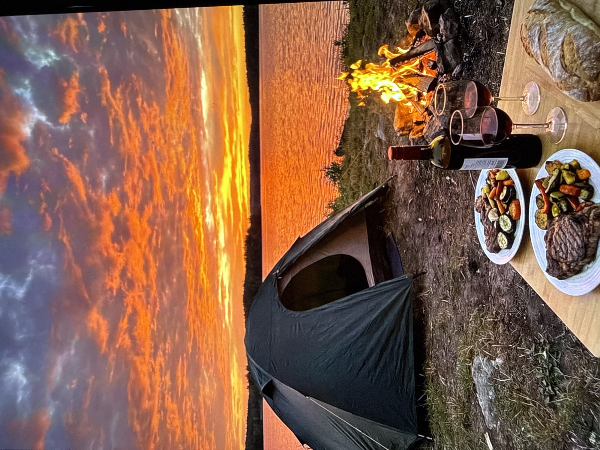 A camping scene is set by a lake during a vibrant sunset. The sky is ablaze with fiery orange, yellow, and pink hues, reflecting on the calm water. In the foreground, a dark green tent is pitched on the grassy bank. A crackling campfire burns to the right of the tent, casting a warm glow.
On a wooden table in front of the campfire, there are two plates of grilled steak and vegetables, a bottle of red wine, and two glasses filled with red wine. A loaf of bread lies to the right of the plates. The overall atmosphere is one of peaceful, rustic dining in a natural setting. The image does not contain any information to suggest it is located in the city of Varna, Bulgaria.