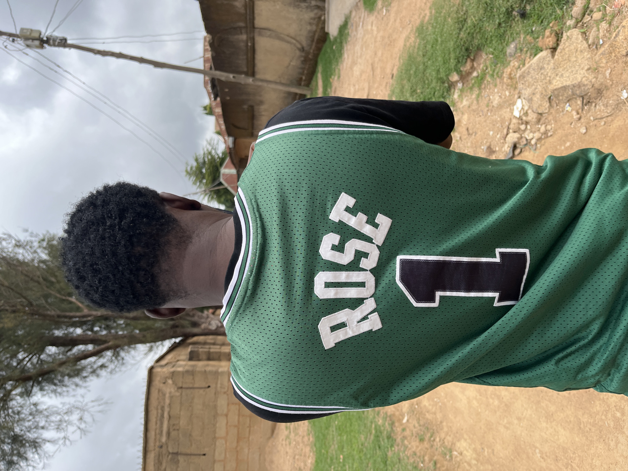 An outdoor, eye-level photograph captures a person from behind, seemingly walking away or standing with their back to the camera. The individual, who appears to be male, has short, dark, curly hair and is wearing a green and black basketball jersey. The back of the jersey features the name "ROSE" and the number "1" in prominent white block lettering with black outlines, set against the green mesh fabric.
The scene depicts a modest, possibly residential or semi-urban setting, likely an unpaved street or path. The ground is a mix of reddish-brown dirt and patches of green grass. In the background, under an overcast sky, there's a utility pole with numerous wires stretching horizontally across the upper frame. Weathered structures, possibly buildings with concrete or brick walls and simple rooftops, are visible in the mid-ground. Sparse trees with some foliage are also present, suggesting an environment with natural elements alongside human infrastructure. The overall impression is of a cloudy day in Barakin Akawo, Nigeria. No discernible emotions are conveyed as only the back of the person is visible.