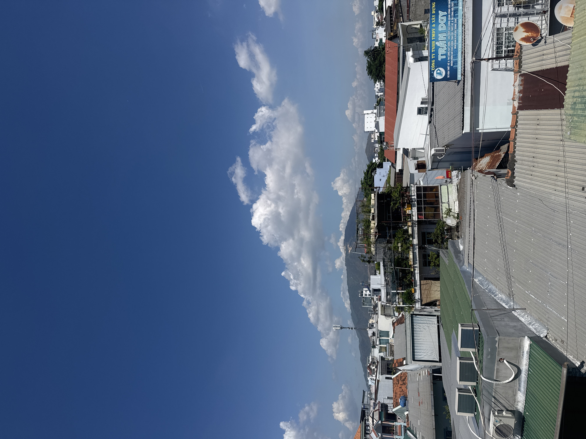 This image shows a cityscape in Nha Trang Ward, Vietnam. The view is from a rooftop, overlooking a dense urban area with various buildings of different heights and colors, primarily white and gray, with corrugated metal roofs. A mountain range is visible in the background. The sky is mostly blue with scattered white clouds. A sign in Vietnamese advertises architectural and construction services. There are numerous air conditioning units, satellite dishes, and other rooftop installations.
