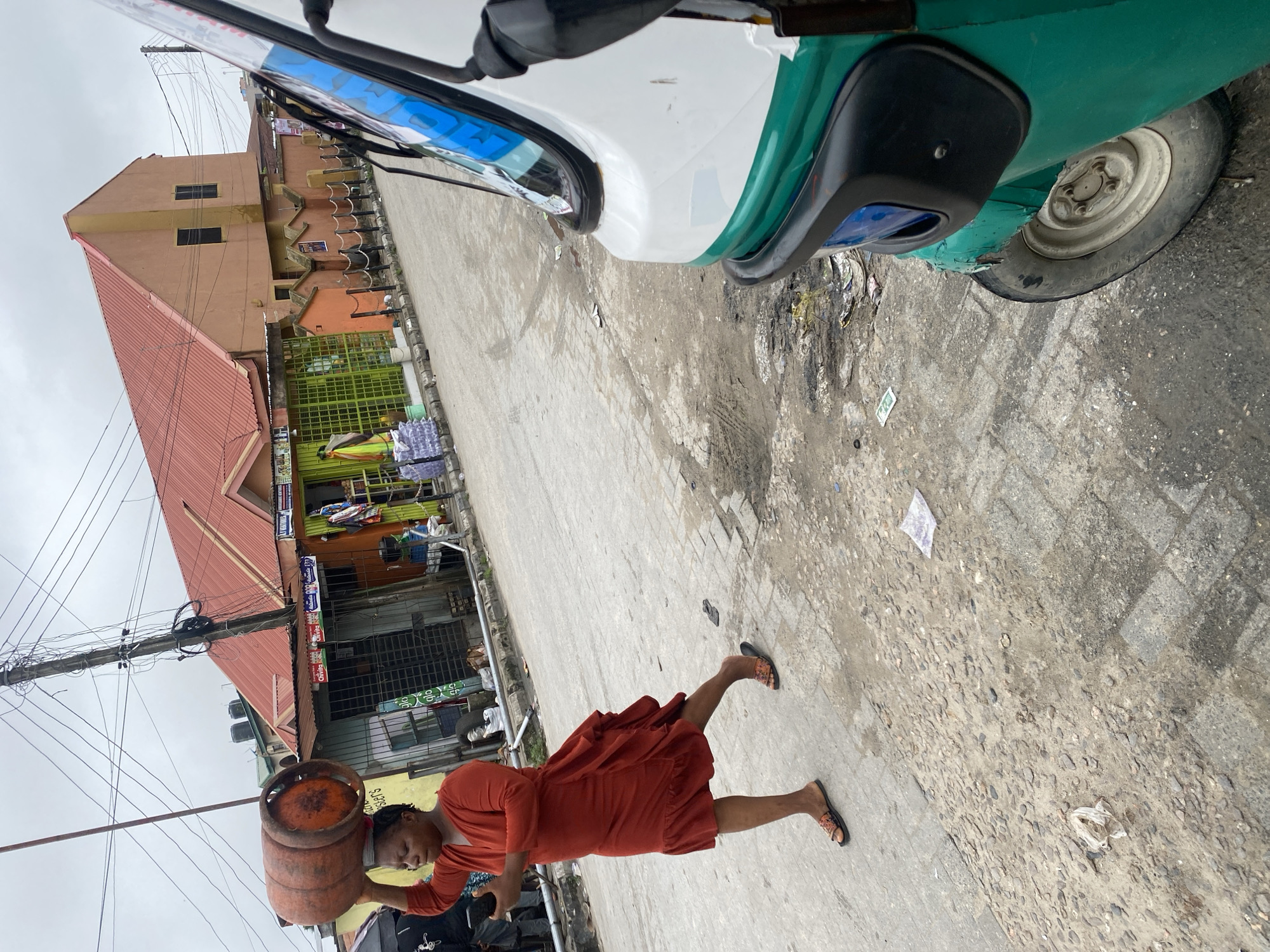 This image depicts an outdoor street scene in Ado, Nigeria, under an overcast sky. In the foreground-left, a woman walks from right to left, carrying a large, orange, rusted LPG cylinder balanced on her head. She is wearing a long-sleeved, rust-colored dress with ruffled detailing on the skirt, and open-toed sandals. Her right hand holds a smartphone, towards which her gaze is directed.
Partially visible in the foreground-right is the front section of a green, white, and blue auto-rickshaw, with its front wheel and headlight assembly evident. The vehicle shows signs of wear.
The background features a row of buildings lining the right side of the street. A prominent two-story building has a reddish corrugated metal roof and a peach-colored facade with two narrow, dark windows on its upper section. Below this, various single-story commercial establishments are visible, including one with a green metal gate and another with pale blue walls. Advertisements for "Chivita" are affixed to some shopfronts. Stacks of bottled water are visible outside one of the shops. A low wall or fence with chained bollards runs in front of some buildings. Overhead, a dense cluster of electrical wires and cables emanates from a utility pole near the center of the image.
The street surface is uneven, consisting of a mix of worn pavement, exposed aggregate, and dirt patches with visible tire tracks. Scattered litter, including plastic bags and paper, is present on the ground. Water tanks are visible on rooftops in the distant background.