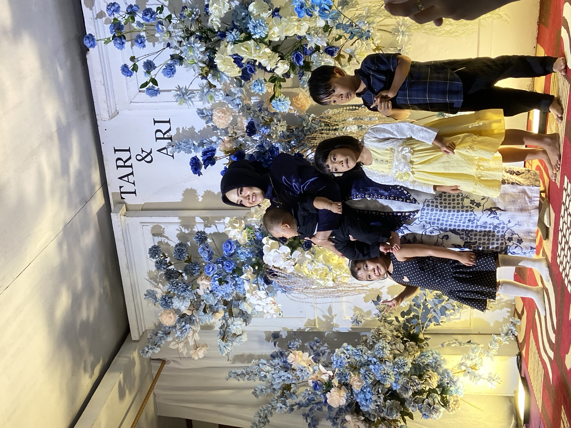 A wedding reception backdrop featuring the names "TARI & ARI" is adorned with large floral arrangements in shades of blue, white, and cream. In front of the backdrop, a woman wearing a dark hijab and a dark blue dress holds a baby. To her left, a young girl in a black polka dot dress and white socks stands with her right arm raised, making a peace sign. To the woman's right, a young girl in a yellow dress and white cardigan stands, followed by a young boy in a blue plaid shirt and black pants. They are positioned on a red and white striped carpeted stage. The scene appears to be indoors, possibly at a venue in Kembang Harum, Indonesia, indicated by a decorative element in the background that suggests a traditional Indonesian wedding setting. Lighting casts shadows on the ceiling, and some of the floral decorations are illuminated.