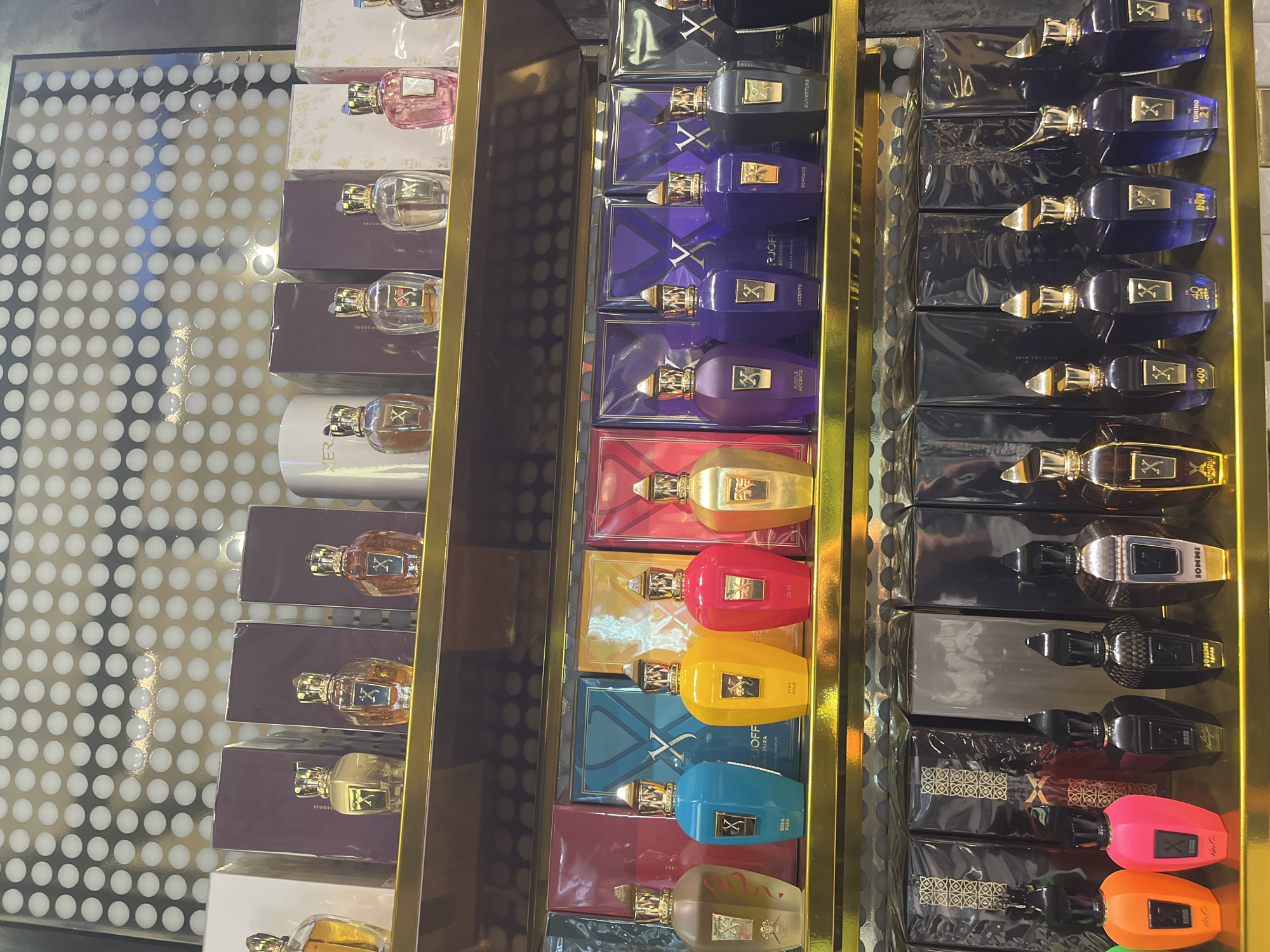 The image shows a shelf display of Xerjoff brand perfumes. The perfumes are arranged in three columns, with the first column containing perfumes in various light shades and boxes, the second column containing perfumes in different colors and boxes with Xerjoff branding, and the third column containing perfumes in darker shades and boxes. The location is unknown and there is no information about people, actions, or interactions. The overall environment appears to be a retail setting.