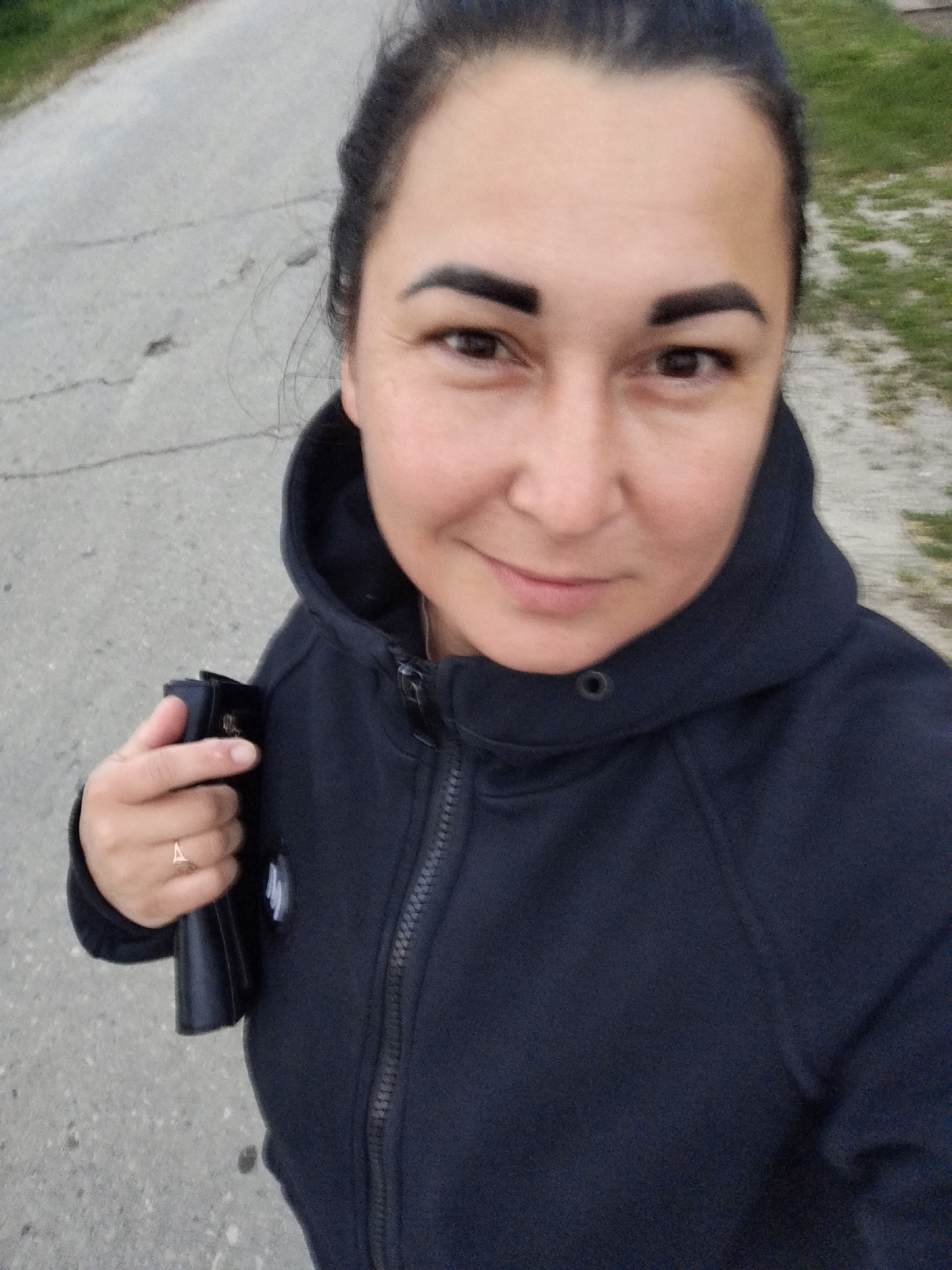 In Krasny Gulyai, Russia, a woman with dark hair pulled back is taking a selfie outdoors. She is wearing a dark hooded jacket with a zipper. In her left hand, she holds a black wallet or clutch. A ring with a triangular design is visible on her ring finger. The background shows a paved road with grass and vegetation alongside it.