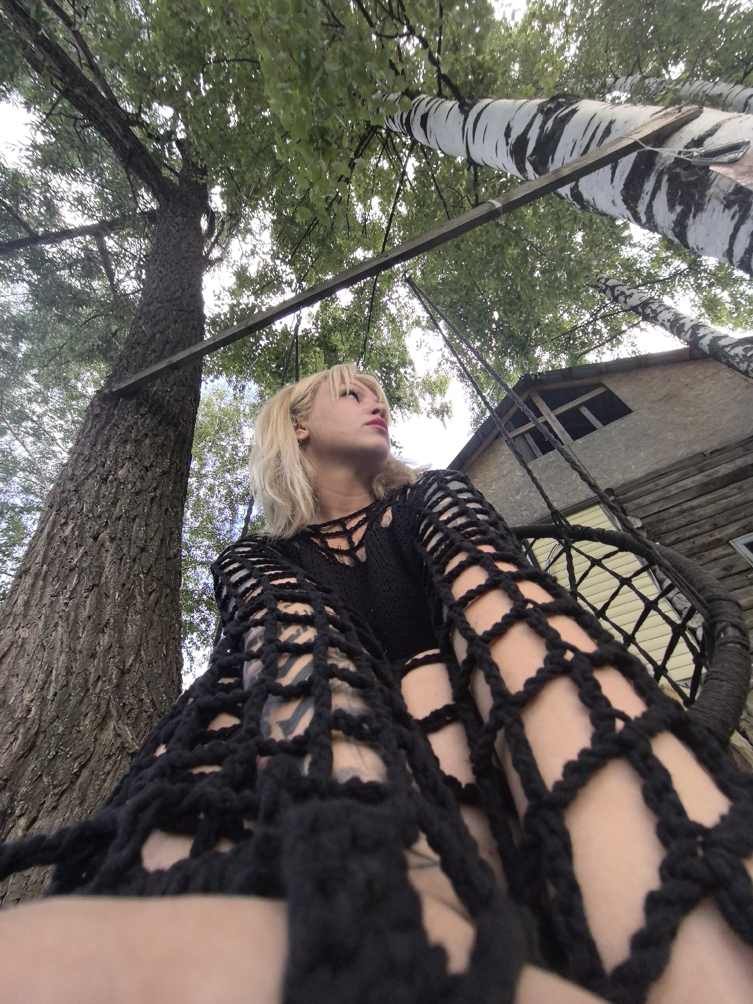 In Noviy, Russia, a blonde woman with light skin, wearing a black crocheted top, is photographed from a low angle while sitting in a hanging chair. She is looking upwards and to the right, with her lips slightly parted. The background features a large tree trunk to the left and a birch tree trunk to the right, with green foliage overhead. A wooden structure, possibly a small house or treehouse, is visible in the background. The scene appears to be outdoors, with natural lighting.