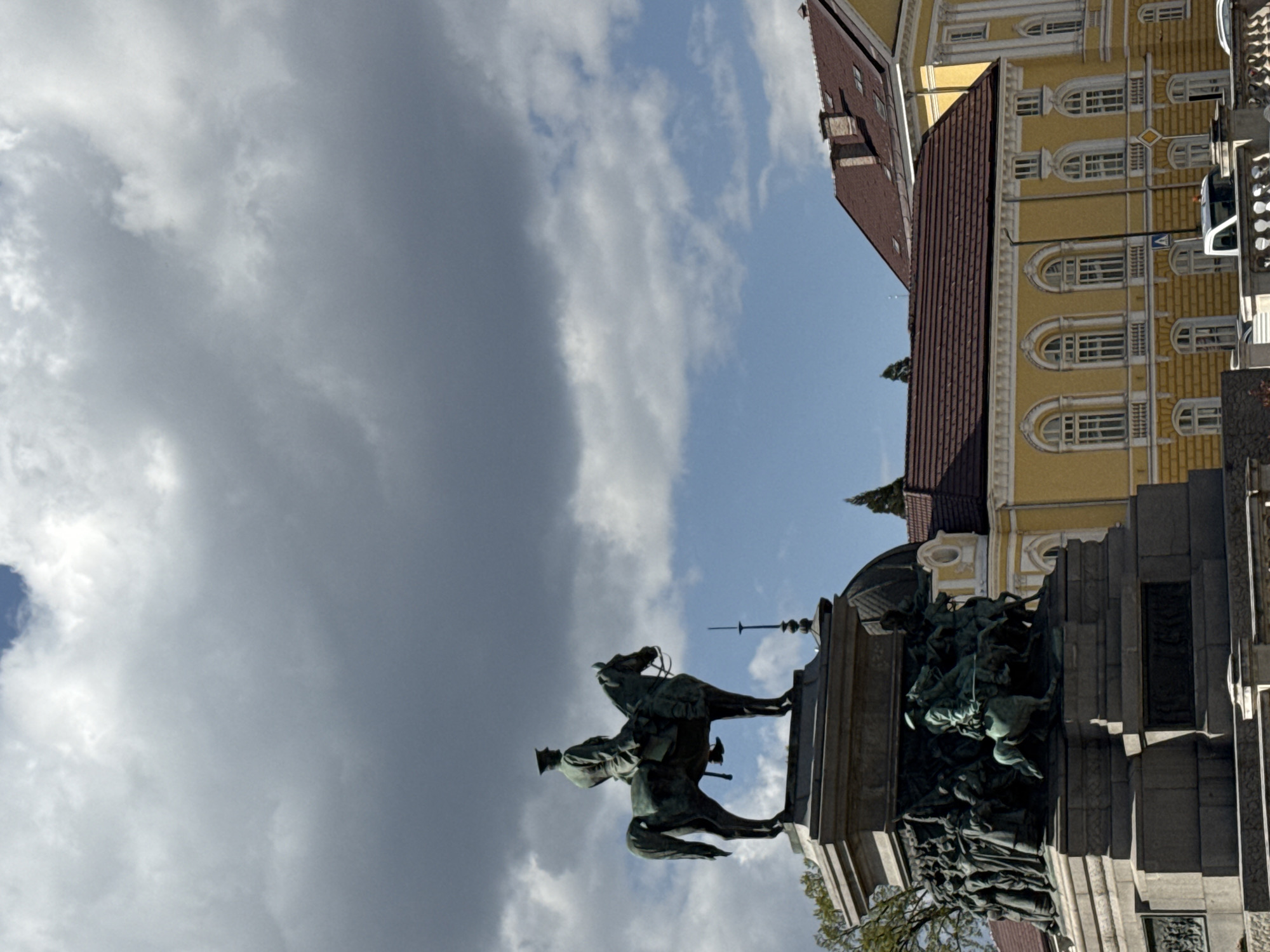 The image, taken in Sofia, Bulgaria, captures an outdoor daytime scene featuring a prominent equestrian monument positioned in front of a historic building, all set under a partly cloudy sky. The central focus is a large, dark-colored equestrian statue depicting a male figure, likely a military leader, in uniform and a hat, riding a dynamically posed horse. This statue rests on an elaborate, multi-tiered stone pedestal. A detailed relief sculpture, showing multiple figures, possibly in a battle or historical event, adorns one side of the pedestal.
To the right and slightly behind the monument, a grand, multi-story building is visible. It features distinctive yellow facades with ornate white architectural details, including numerous arched windows, and is topped with a reddish-brown tiled roof. Another building with a similar roofline is partially seen further left. A white vehicle, possibly a van or small truck, is partly visible near the yellow building, on which a yellow diamond-shaped road sign with a black 'X' is affixed.
The sky occupies the upper and left portions of the image, displaying a mix of large, grey clouds and patches of clear blue. Green foliage from trees can be seen near the base of the monument and around the buildings. The bright, natural illumination suggests a daytime setting, possibly late morning or early afternoon. The architectural style and the monument are characteristic of a historic European capital. No discernible text is clearly visible on the monument or buildings.