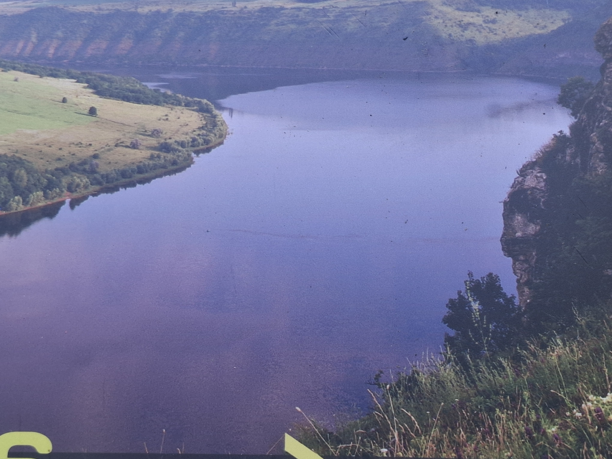 The image depicts a wide river winding through a valley with steep, forested slopes. The water is a deep blue, reflecting the sky and surrounding landscape. The foreground shows a grassy cliffside with wildflowers and tall grasses. In the distance, rolling hills covered in green vegetation recede into a hazy horizon. There are no people or obvious man-made structures visible in the natural landscape. The overall impression is one of a serene, remote natural setting. It is difficult to determine the exact location within Vadul lui Voda, Moldova, without further context or identifiable landmarks.
