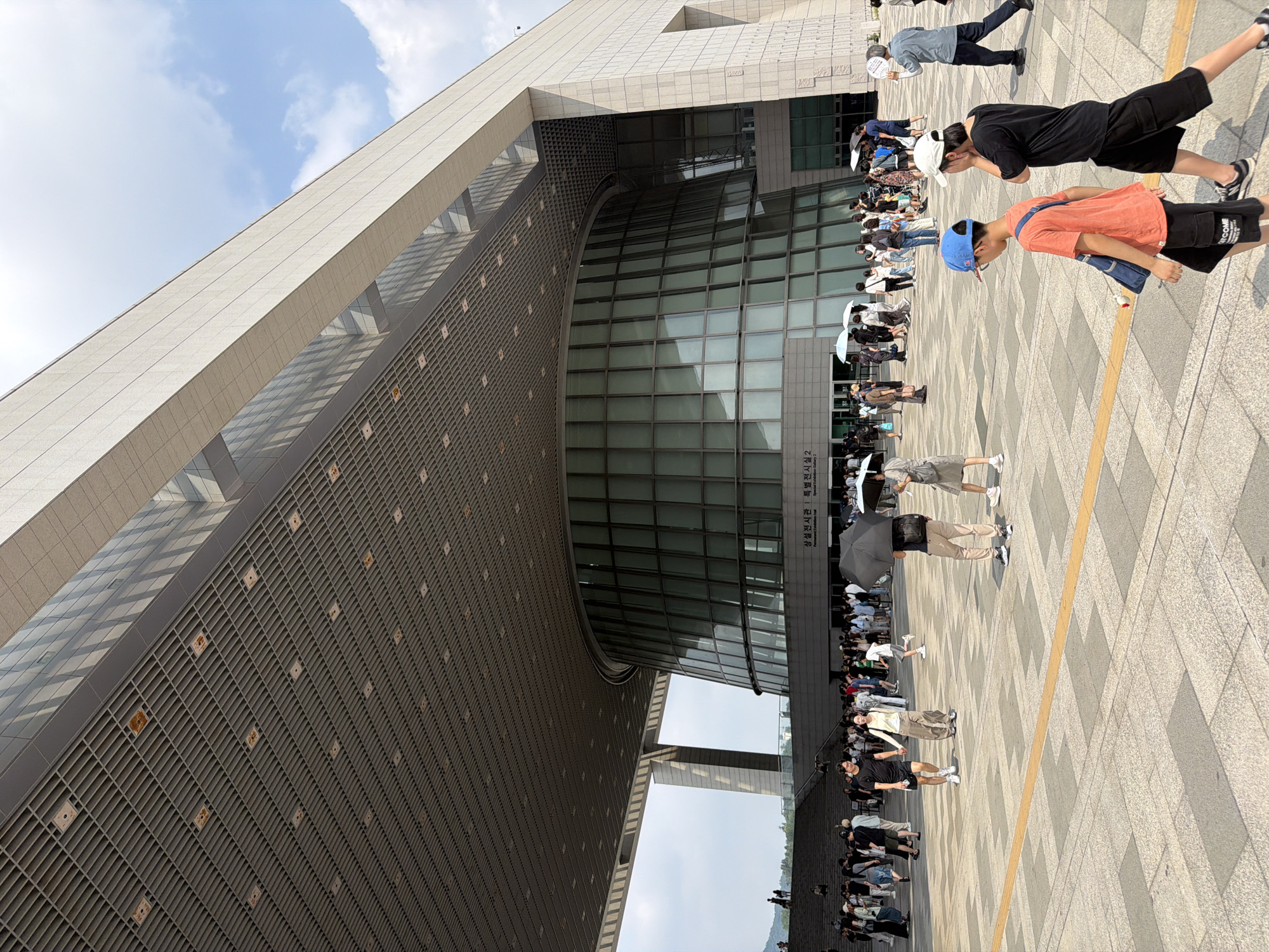 This image depicts the exterior of the National Museum of Korea, located in Yongsan-gu, Seoul, South Korea. The foreground shows a paved plaza with several individuals walking. In the middle ground, a large, circular glass-fronted building, part of the museum complex, is visible, along with a group of people gathered near its entrance. Overhead, a massive, grey, latticed canopy extends from the museum, casting a shadow over the plaza. The background features a bright, partially cloudy sky. People are dressed in casual summer attire, and some are holding umbrellas, suggesting warm or potentially rainy weather.