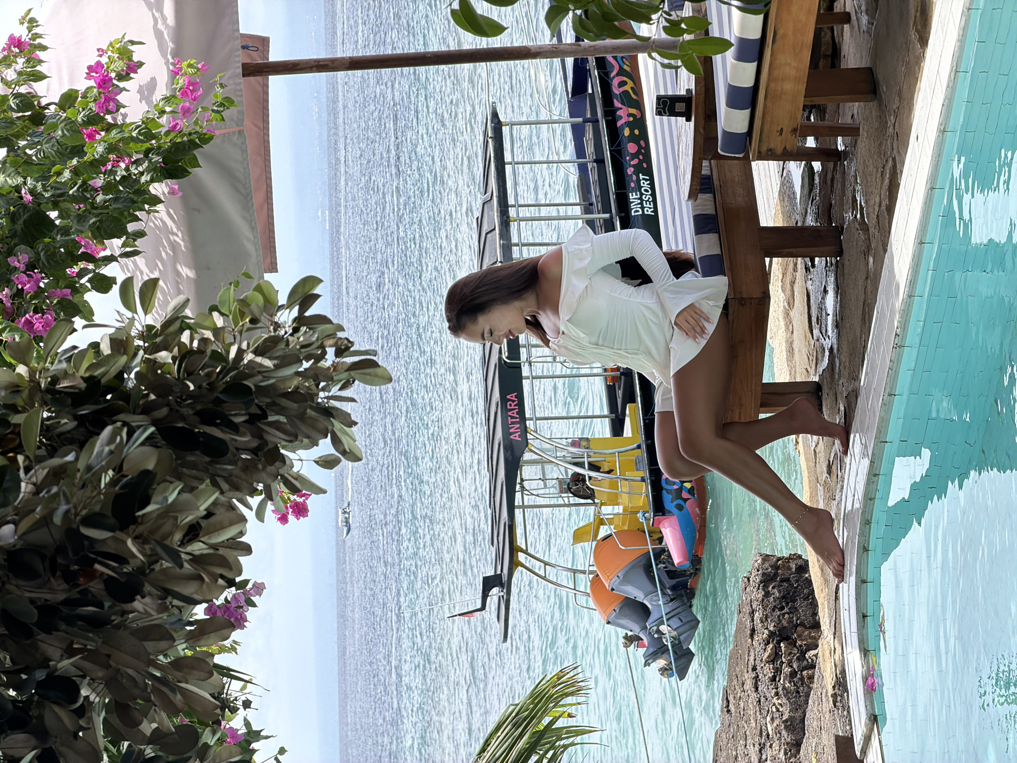 The image captures a woman in a white dress near a poolside setting in Nusa Penida, Indonesia. She leans against a wooden structure, and her feet are near the edge of a swimming pool. In the background, boats are visible on the water, including one labeled "ANTARA DIVE RESORT." Lush greenery with flowering plants frames the scene, adding to the coastal ambiance.