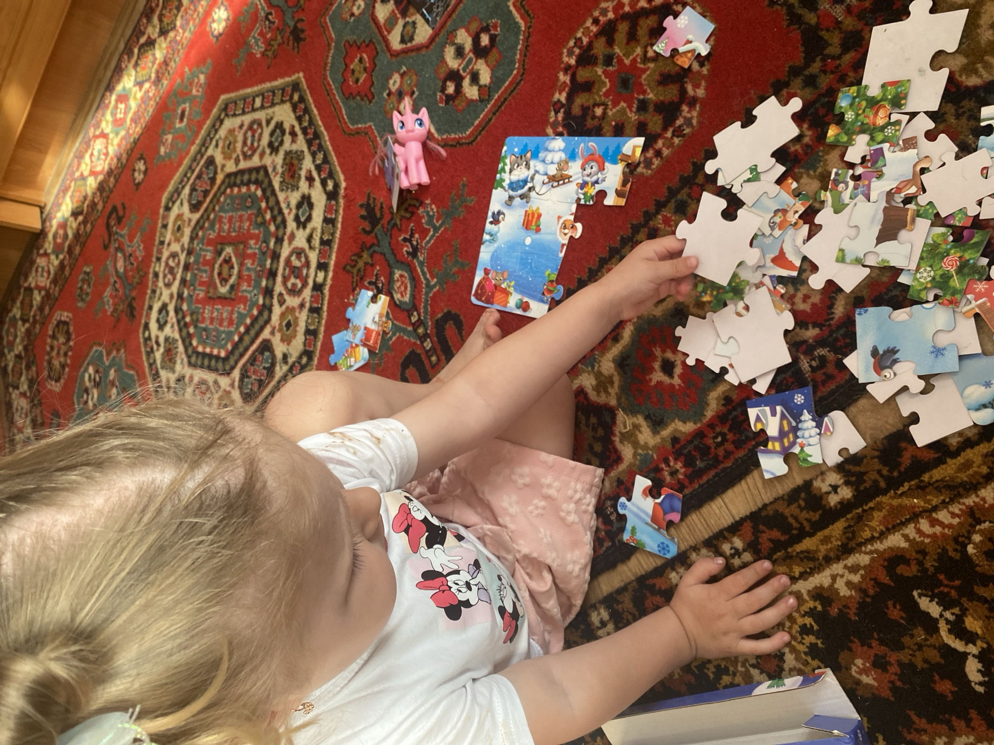 A blonde-haired toddler, wearing a white t-shirt with Minnie Mouse graphics and a pink floral skirt, sits on a red oriental rug and plays with a jigsaw puzzle. The puzzle depicts various characters, including cats, rabbits, and a fox, set against a wintery background with snow-covered houses and Christmas trees. Several puzzle pieces are scattered around the child, some assembled and some not. A pink toy pony is visible in the upper right corner of the frame. The scene is captured from a high angle, looking down at the child and the puzzle. The environment suggests an indoor setting, possibly a living room or playroom, with wooden furniture visible in the background. The image does not provide any information about the city of Маринопіль, Ukraine, or any specific location details.