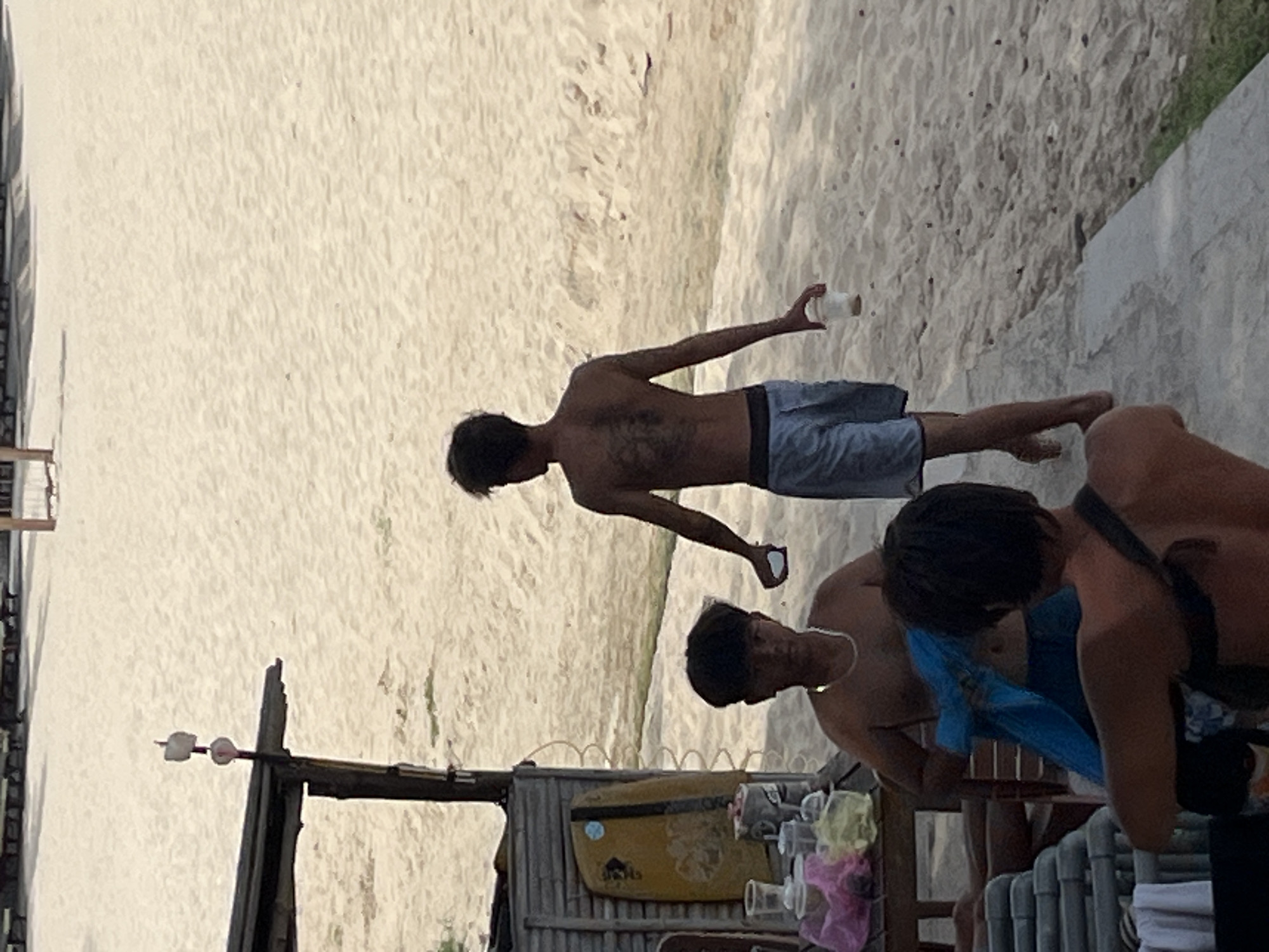 The image depicts a beach scene in Phu Thuy Ward, Vietnam, with a focus on three individuals and their surroundings. In the foreground, a person is partially visible from the back, wearing a dark-colored strap across their shoulders. To their left, another person is seated, wearing a necklace and holding a blue fabric. Further back, a third individual is walking away from the camera, facing towards the beach, wearing light blue shorts and holding a white cup in their right hand. This individual has a prominent tattoo on their back.
The environment consists of a sandy beach with some sparse vegetation and a wooden structure to the left, which appears to be a stall or shelter. This structure has a yellow surfboard leaning against it and several plastic cups and bags on a wooden table. In the background, rows of empty lounge chairs are visible, suggesting a recreational area. The overall lighting indicates it is daytime, likely late afternoon, given the soft shadows.