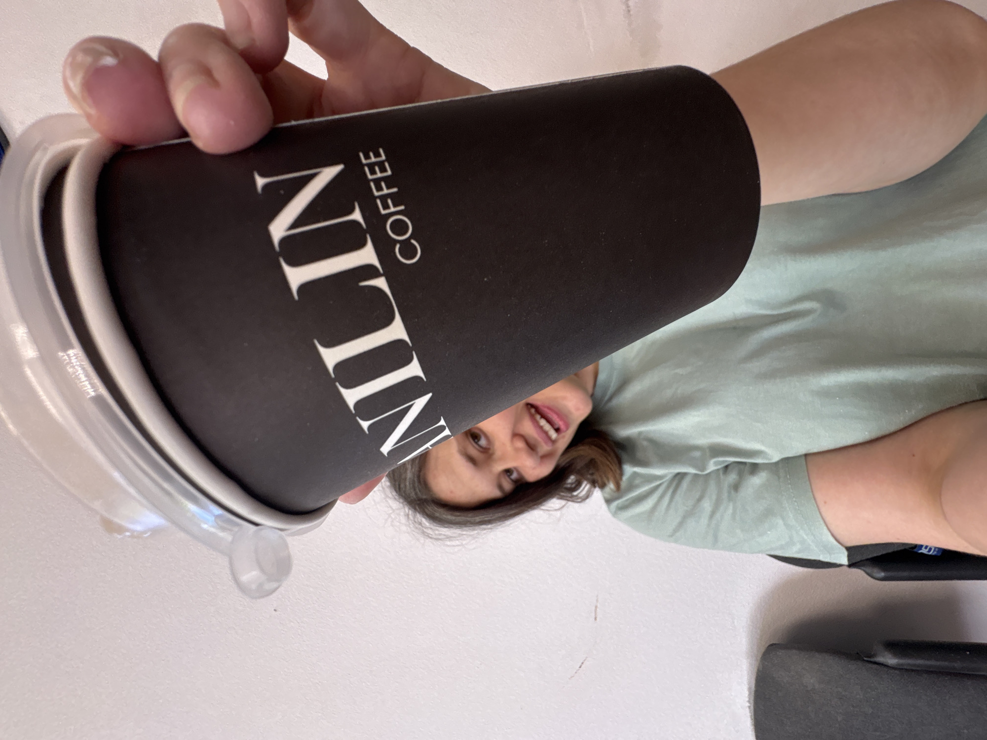 A woman holds a black coffee cup with white "Nulin Coffee" text. The cup has a clear plastic lid. The woman has short brown hair and is wearing a light green shirt. The background consists of a white wall and a black object. The location is Chaikovsky, Russia.