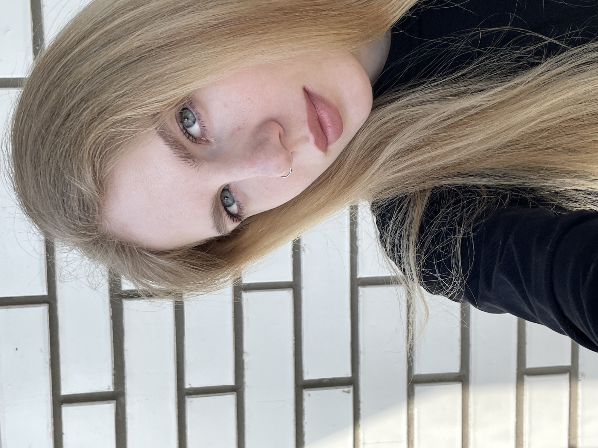 A close-up portrait shows a young woman with fair skin, light blonde hair, and green eyes. She has a nose ring and is wearing a dark-colored garment. The background is a white brick wall. The location is not discernable as Izhevsk, Russia, from the details within the image.