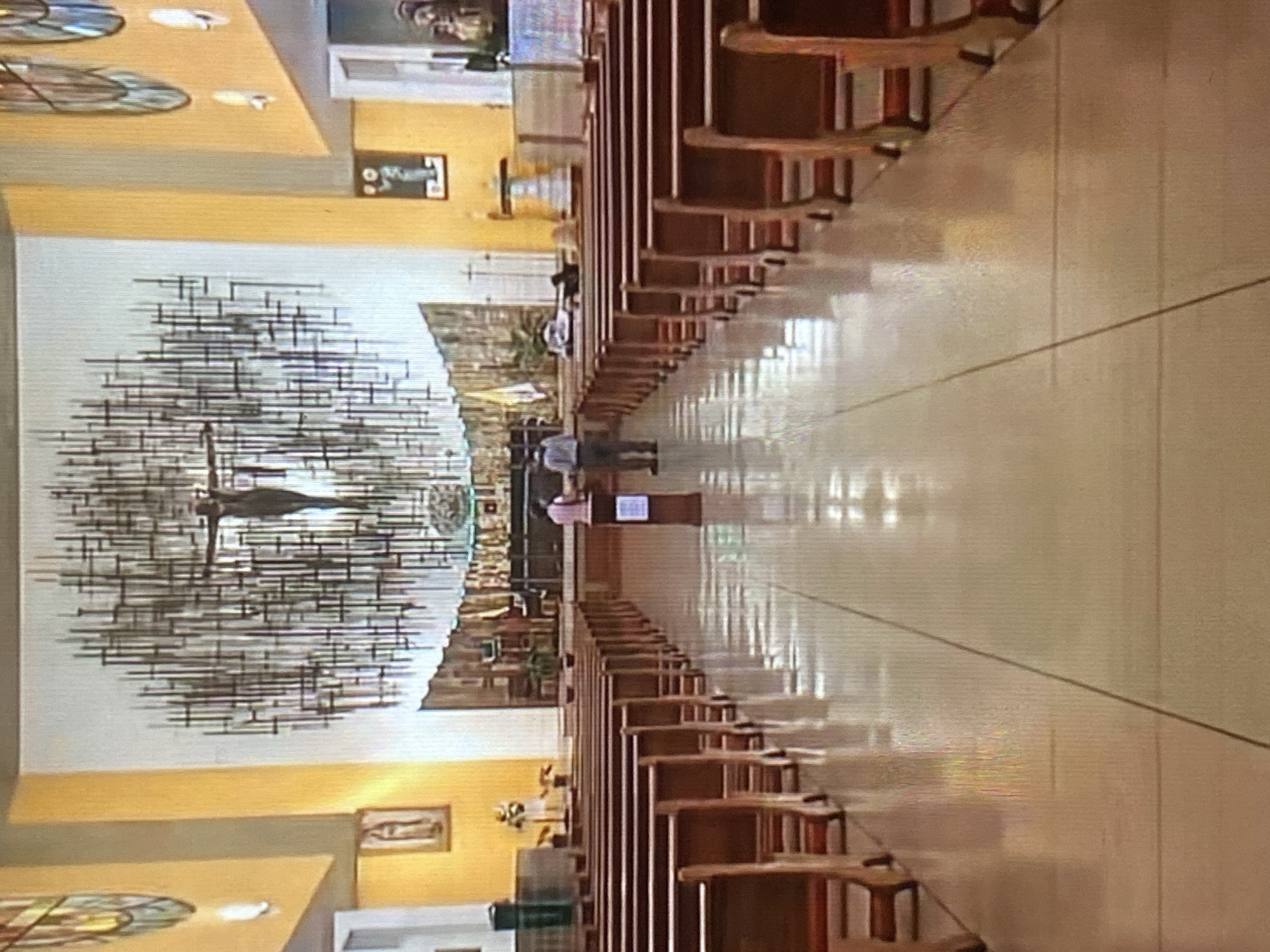 This image captures the interior of a church or chapel, looking towards the altar from the back rows of pews. The image is rotated clockwise, so the pews appear to extend upwards along what would be the floor.
In the foreground and extending towards the front, rows of dark wooden pews with lighter seats are neatly arranged on both sides of a central aisle. The floor of the aisle is reflective, showing distorted reflections of the pews and ceiling lights, indicating a polished or tiled surface.
At the front of the church, on the main wall, a prominent and dramatic metal sculpture of a crucified Christ figure is the focal point. The sculpture features a central figure surrounded by numerous horizontal and vertical metallic lines, creating a striking and abstract visual effect. Below this large sculpture, another, less distinct artwork or mural can be seen.
Two individuals are visible standing in the central aisle near the front, between the pews, facing the altar. One person, wearing a light-colored top and dark trousers, is more clearly visible than the other. A small stand or podium-like object is also present in the aisle near them.
Along the upper parts of the side walls, stained-glass windows are visible, allowing light into the space. Additional framed artworks or pictures adorn the side walls. Recessed lighting fixtures are visible on the ceiling and side structures, supplementing the natural light.
The setting suggests a quiet moment, possibly before or after a service, or during a private visit for prayer or contemplation. The overall atmosphere is one of reverence within a structured religious space. The location context is Tinaquillo, Venezuela.