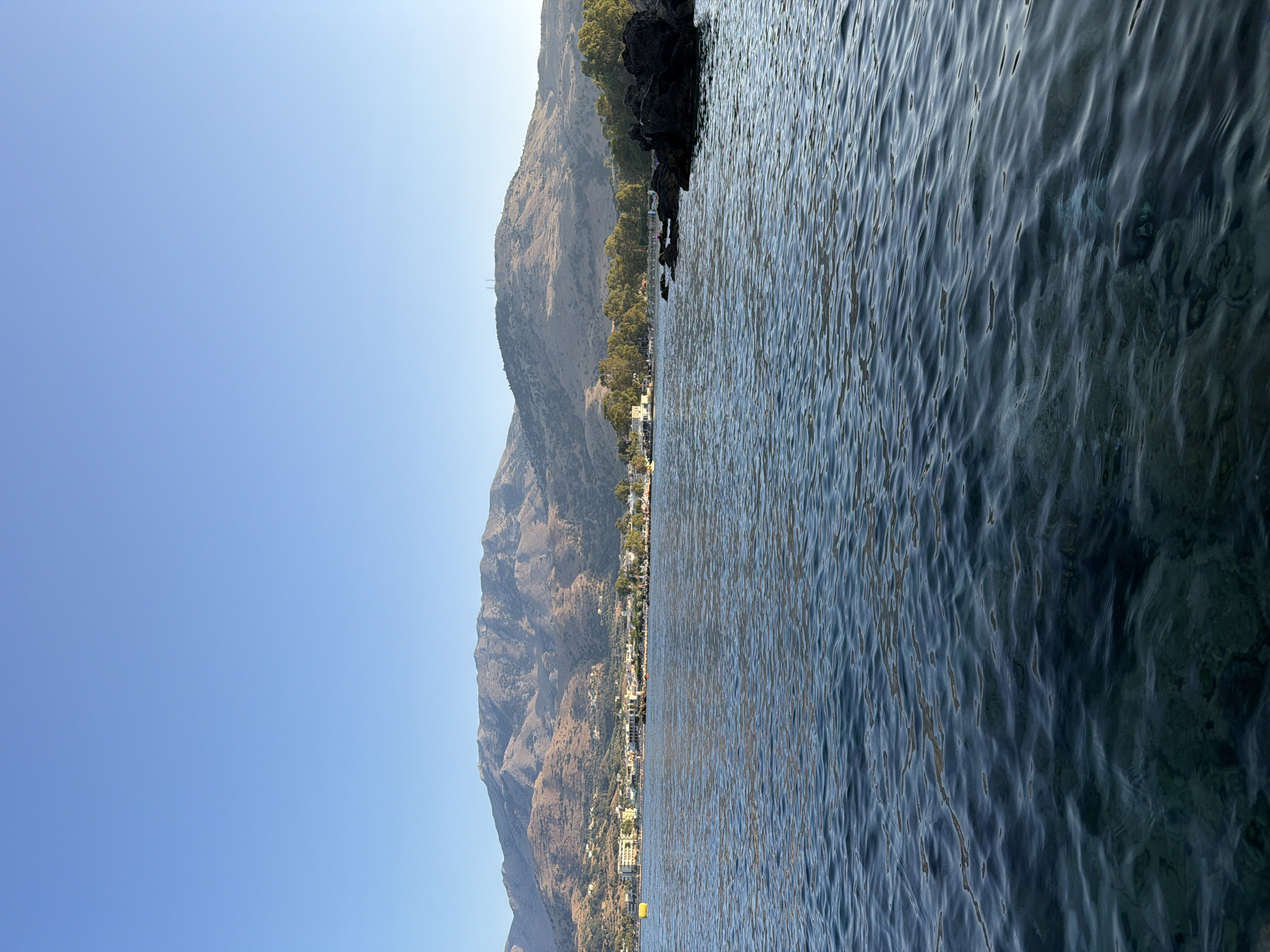 A wide, clear blue sky stretches over a tranquil blue sea, its surface rippled by a gentle breeze. In the distance, a coastal town nestled at the foot of a majestic mountain range comes into view. Buildings, primarily in light colors, form a visible line along the coast, interspersed with scattered trees. The mountains themselves are rugged and bear the marks of time and weather, their slopes a mosaic of earthy tones. To the right of the frame, dark, jagged rocks emerge from the water, some with a few people visible nearby. A solitary yellow buoy floats in the sea further to the left, marking its presence against the vast expanse of water. The scene evokes a sense of serene coastal beauty, characteristic of a location like Georgioupoli, Greece.