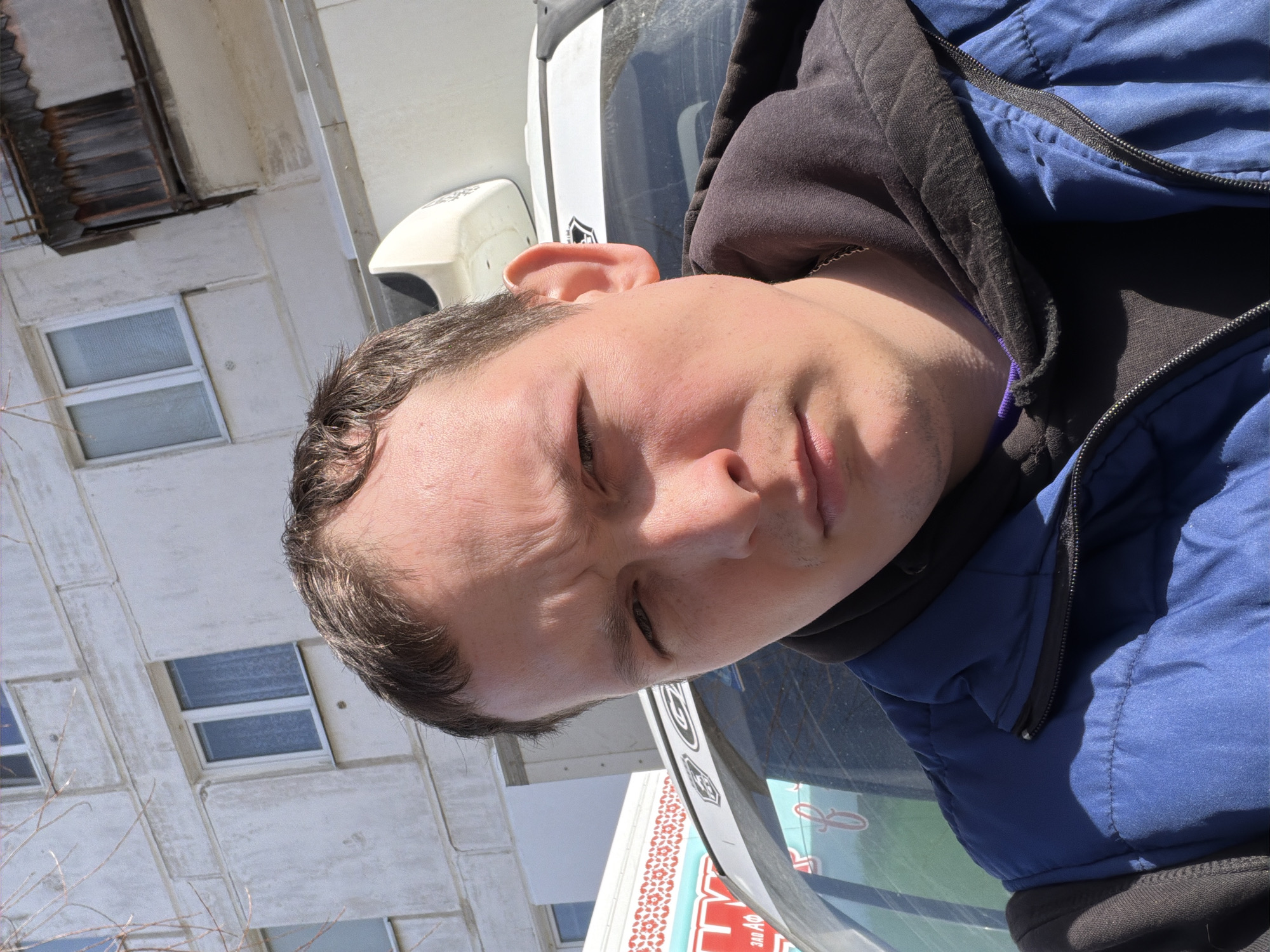 The image shows a close-up, selfie-style shot of a man with short brown hair, wearing a dark hooded sweatshirt underneath a blue puffer jacket. He appears to be standing near a vehicle, possibly a truck, and the reflection of a building and a sign are visible in a windshield. The building is a concrete structure with several windows. The sign seems to be written in Russian. Based on the sign's content and the architectural style, the location is suspected to be in the city of Chaikovsky, Russia. The man's face displays a neutral or slightly pensive expression. The photo appears to be taken outdoors in daylight.