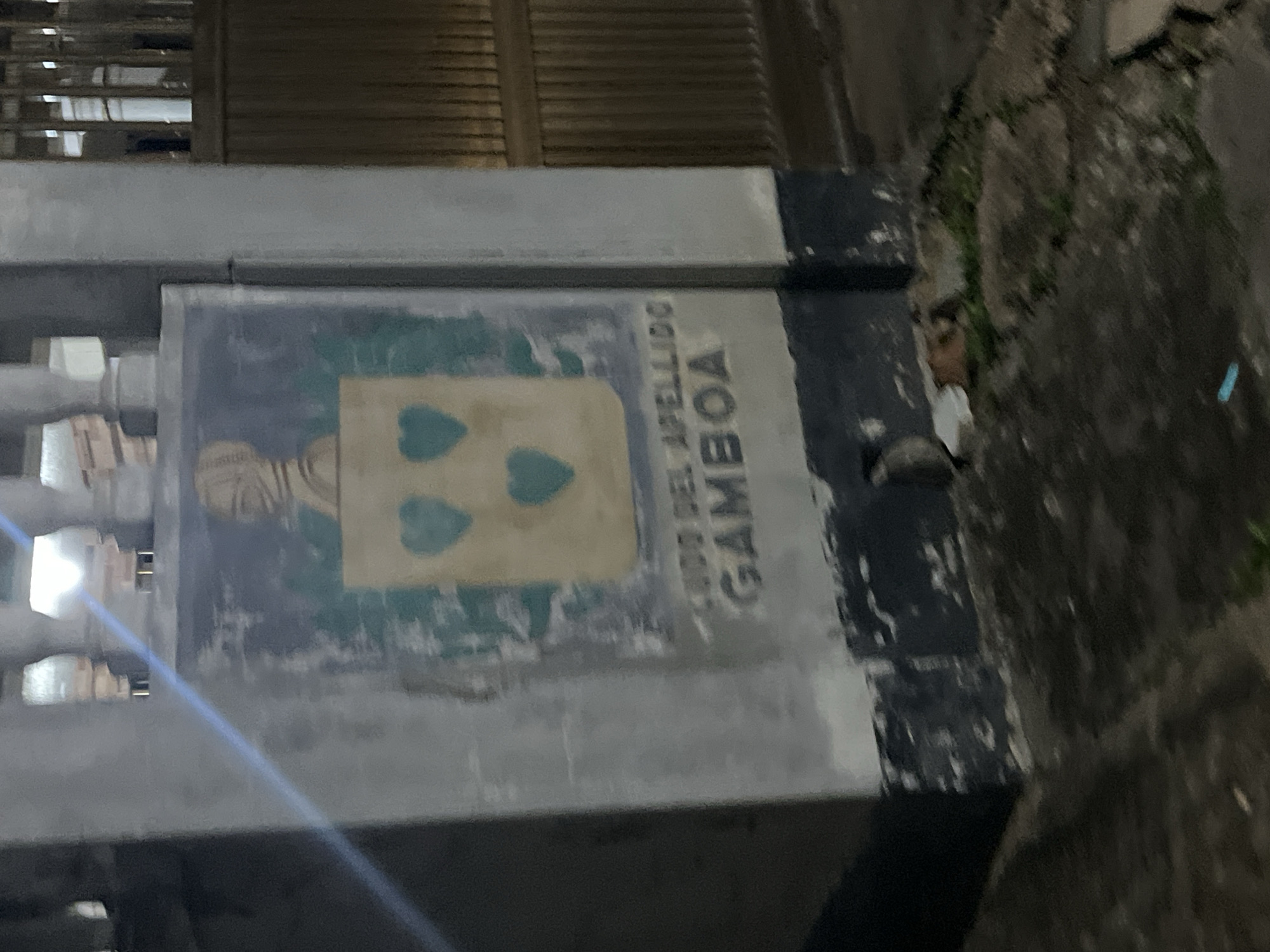 The image captures a portion of a weathered concrete pillar in Santa Ana, Venezuela, featuring a faded coat of arms. The coat of arms is depicted on a light-colored shield with three green hearts arranged in a triangular pattern. Above the shield, there is a stylized representation of a bust, possibly a portrait, wearing a helmet or headdress. Below the shield, text is visible, with the words "APELLIDO" and "GAMBOA" clearly legible, suggesting it may denote a family crest or surname. The pillar itself is painted in various shades of gray and shows signs of age and wear. The base of the pillar is painted with a dark blue-green color. The surrounding environment appears to be an outdoor setting, possibly near a street or entrance, with a corrugated metal fence visible in the background. The ground in front of the pillar is uneven and appears to be a mixture of dirt and possibly some paved surfaces. A small amount of vegetation is visible. The lighting suggests it is either dusk or nighttime, with some artificial light sources creating reflections and affecting the overall clarity of the image.