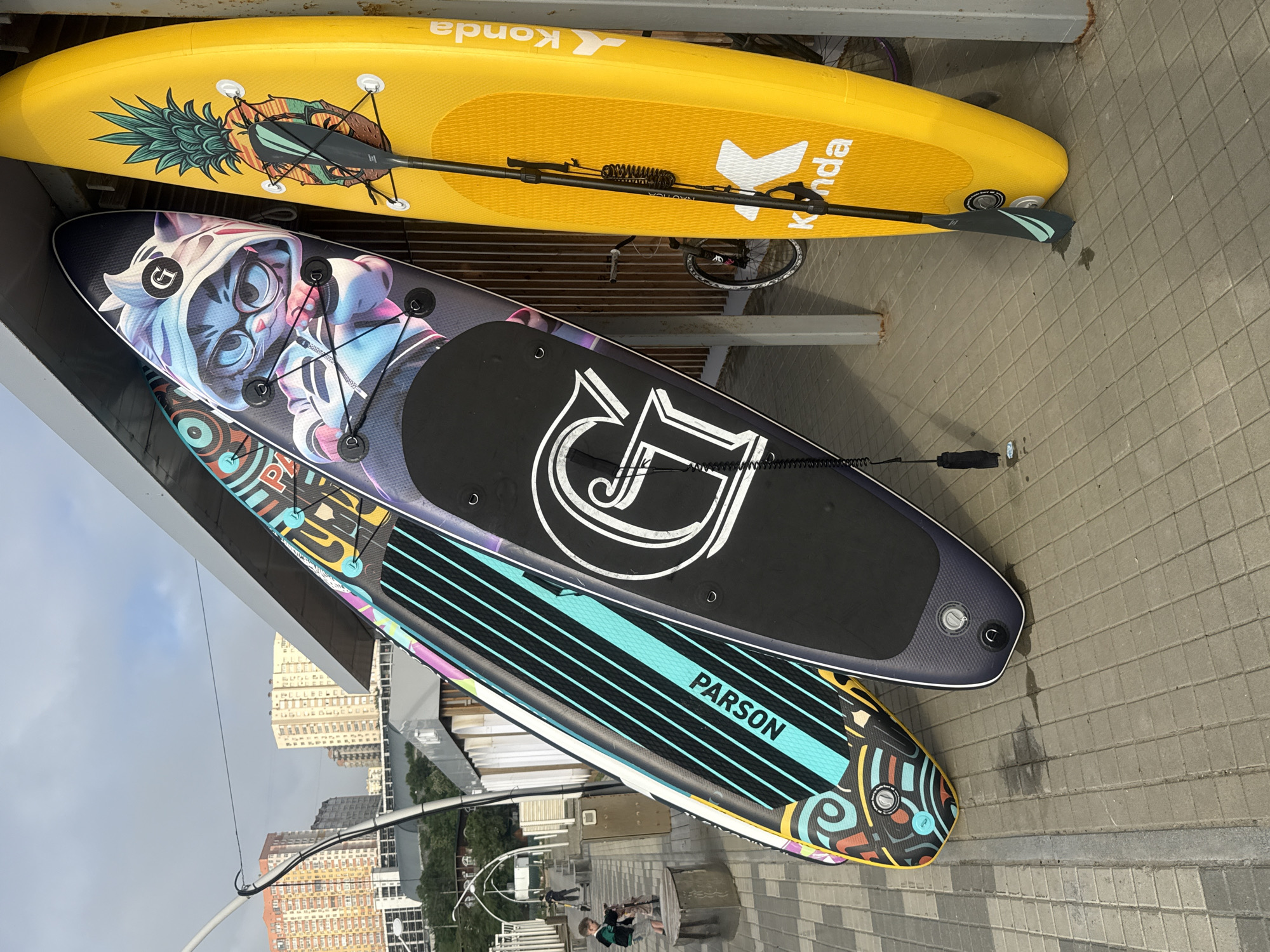 Three paddleboards are propped up against a covered structure with a metal roof and wooden siding. The paddleboards are leaning against each other. The leftmost paddleboard has a colorful abstract design with turquoise and black stripes. The middle paddleboard features an anime-style cat character wearing glasses and a hooded jacket. The rightmost paddleboard is bright yellow and has a pineapple graphic. A paddle is attached to the yellow paddleboard by bungee cords. In the background, there are buildings, a street with a sidewalk, and a cloudy sky. A bicycle is partially visible behind the paddleboards. A young boy is on a scooter in the distance, and another person is sitting on a bench. The scene appears to be a waterfront area. The presence of the "Konda" logo on the yellow paddleboard and the style of the buildings suggest this location may be the waterfront in Vladivostok, Russia, specifically near the sports harbor or a similar recreational area.