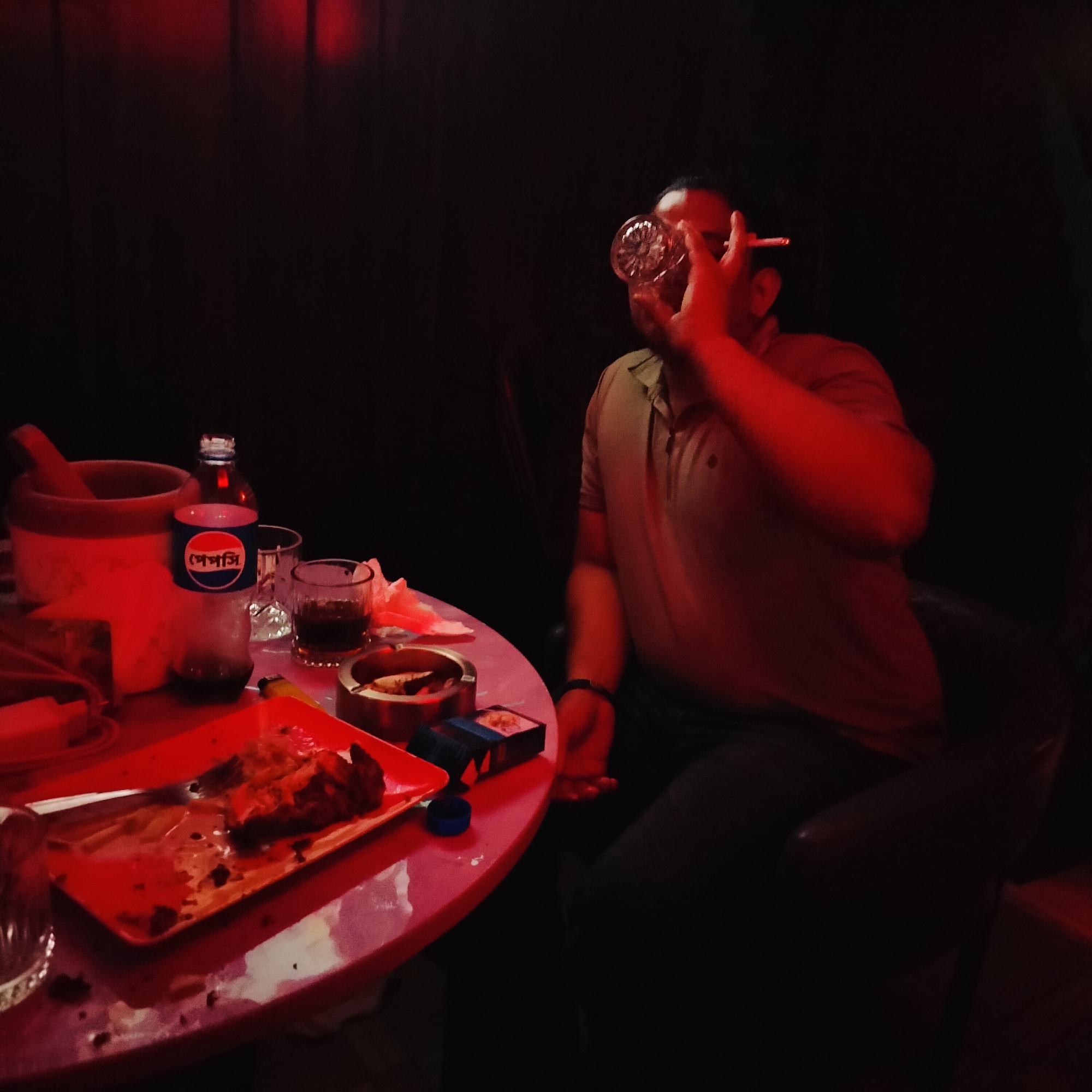 The scene depicts a dimly lit room, possibly a bar or lounge, in Dhaka, Bangladesh. A man is seated in a chair, holding a patterned glass up to his face and smoking a cigarette. A round, red-colored table is in the foreground, cluttered with items including: a plate with food remnants, an ashtray with cigarette butts, a bottle of Pepsi with Bengali script, other glasses, an ice bucket with a pestle, and a pack of cigarettes. The background is dark, suggesting a closed, indoor space with limited lighting. The lighting is primarily red, casting a warm tone over the scene. The atmosphere appears relaxed and casual.