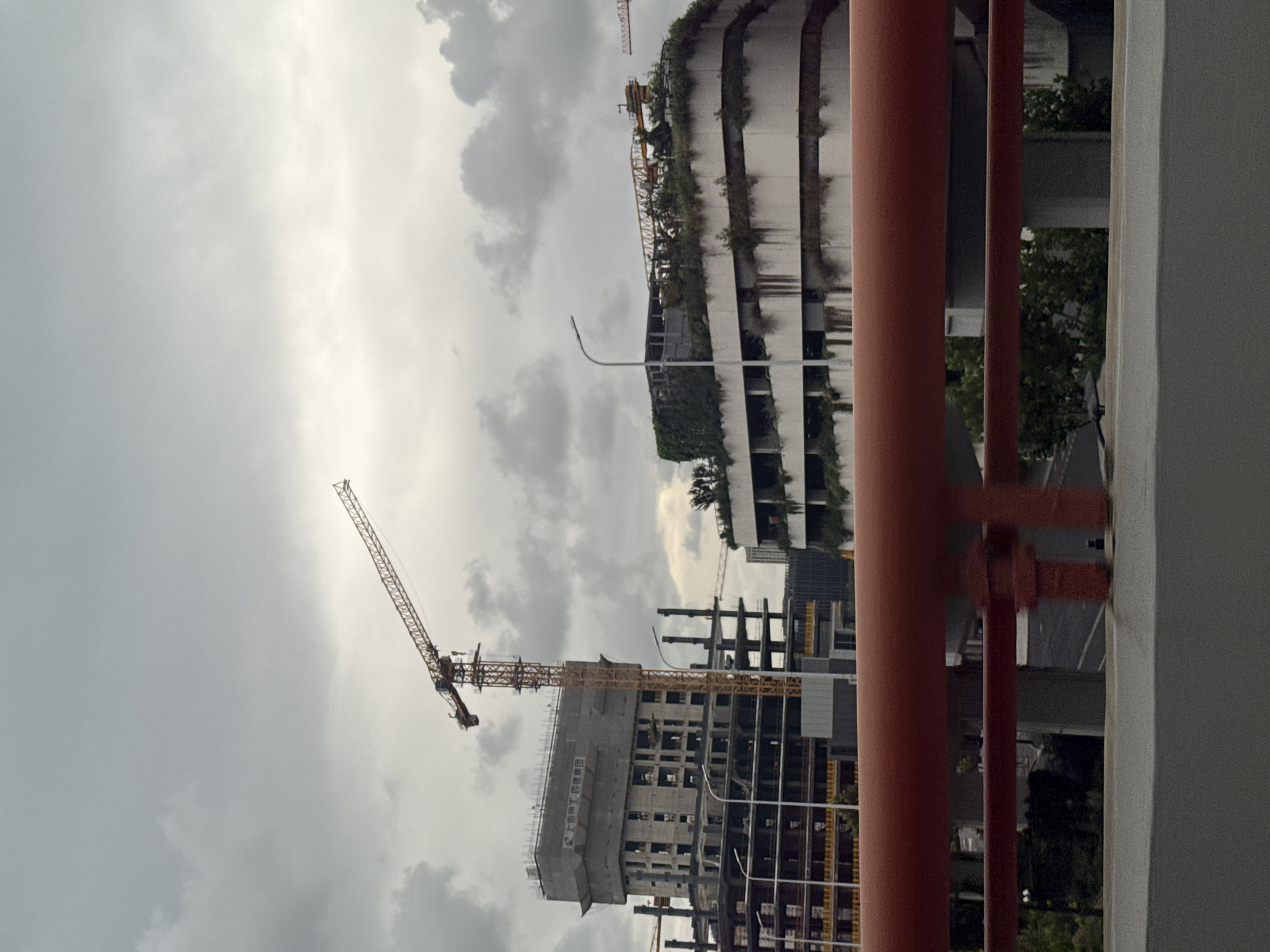 This image captures an overcast sky over a construction site in Shanghai, China. In the foreground, a red metal railing runs horizontally across the frame, obscuring the lower portion of the scene. Behind the railing, several buildings are under various stages of construction. A tall yellow tower crane dominates the left side of the image, its long arm extended over a multi-story building wrapped in gray sheeting. This building appears to be a concrete structure with exposed floors and scaffolding. To the right, a curved, multi-level building with a green, overgrown roof is visible. It also appears to be a concrete structure, with some sections showing signs of wear and tear. The environment suggests an active urban development area, with the cloudy sky indicating a potentially humid or rainy climate. The presence of multiple cranes suggests a significant construction project underway.