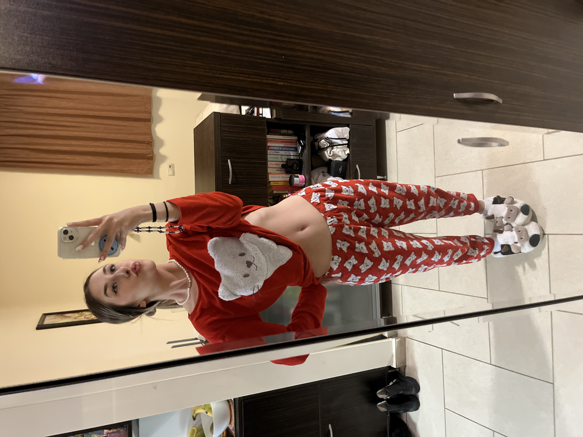 A young woman captures a mirror selfie in an indoor setting, likely a bedroom or living area, in Sofia, Bulgaria. The image is presented rotated 90 degrees counter-clockwise.
The subject is a young woman, visible from approximately the knees up, holding a light-colored smartphone with a blue circular grip to take the picture. She has dark hair, a pearl necklace, and a black hair tie on her left wrist. She is dressed in red long-sleeved pajamas; the top features a white teddy bear appliqué, while the bottoms are patterned with numerous small white teddy bears. Her midriff is exposed between the top and bottoms. She wears white, fluffy slippers shaped like animal faces, possibly sheep or cows. Her expression is a subtle pout, as she looks directly into the phone's camera.
The background, seen in the mirror's reflection, reveals elements of a comfortable home environment. To the left, a window is covered by brown horizontal blinds or curtains. Below, a light-colored wall features an electrical outlet or light switch. To the right, a dark brown wooden cabinet or bookshelf, equipped with silver handles, holds an assortment of books, some with discernible titles like "FORGE", "HEMMINGS", "CAMPUS CRAWL", and "LYLE". Alongside the books are some white or cream fabric items. The floor is covered with large, light-colored square tiles. The attire and indoor setting suggest the photo was taken during the evening or morning.