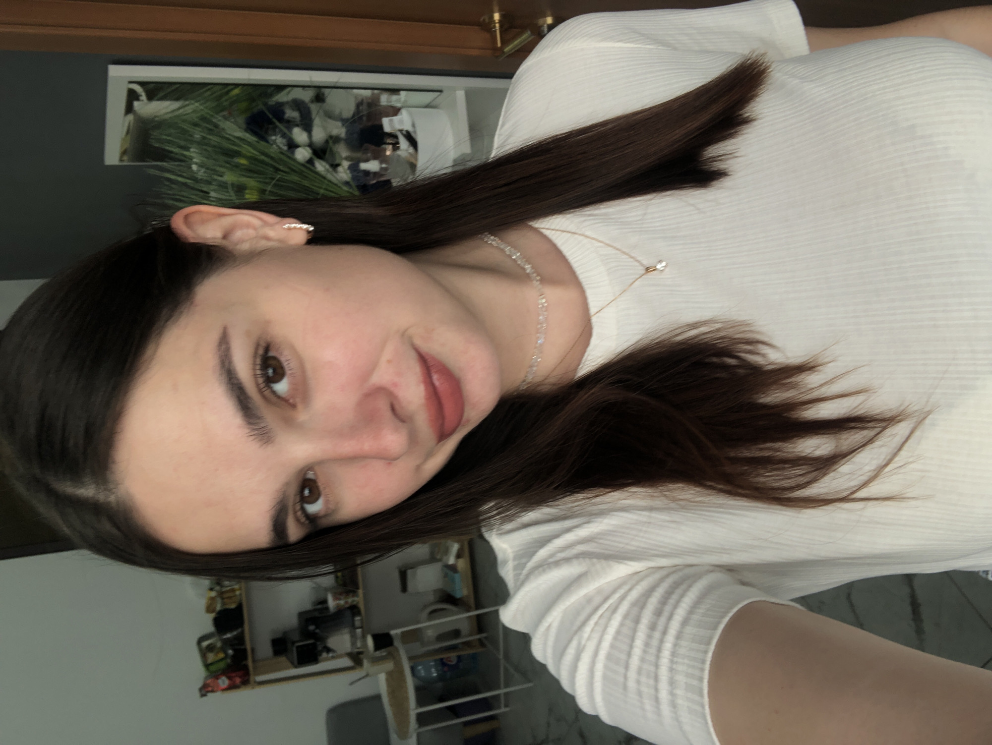 This media file is a close-up, rotated image of a young woman. She has dark brown hair, brown eyes, and is wearing a white, ribbed t-shirt and a delicate necklace. The background appears to be an indoor setting with a dark gray wall and a shelf unit visible in the lower part of the frame, containing various objects. A framed picture with greenery is hanging on the wall behind her. The lighting suggests it is daytime. The woman's expression is neutral, with her head tilted slightly and her gaze directed towards the camera.