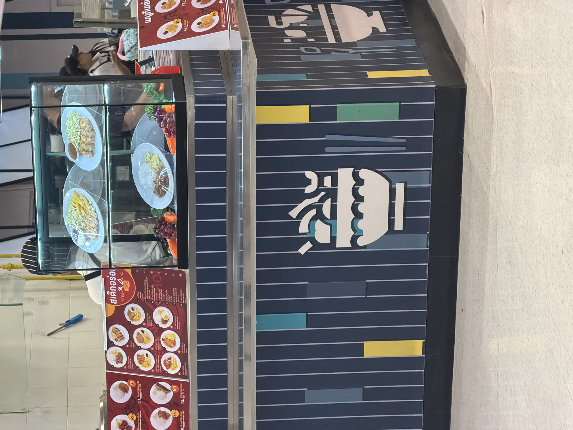 This image depicts a food stall in a marketplace, likely located in Ban Bang Rak, Thailand. The stall features a refrigerated display case showcasing various food items, including plates of rice with meat and vegetables. A menu board with Thai text and prices is prominently displayed, indicating a range of dishes like "Steak Aroi" and various rice-based meals. Two individuals, possibly food vendors, are visible behind the counter, one wearing a chef's hat and the other a baseball cap. The stall itself is decorated with vertical blue and white stripes, accented with colored rectangular blocks and food-related graphics, such as bowls and cutlery. The surrounding environment appears to be an indoor market or food court, with a light-colored floor and neutral walls.